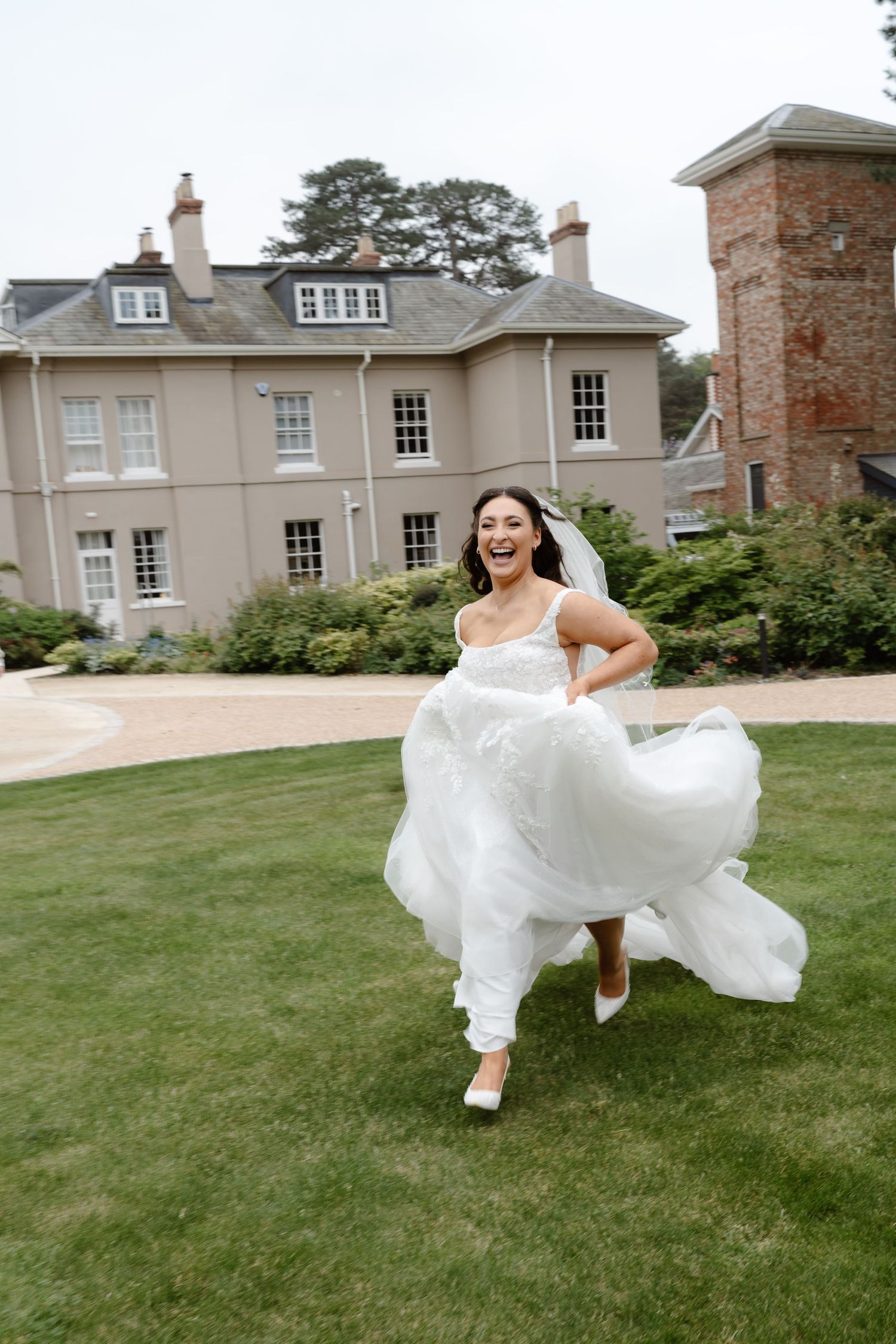 A joyful bride in a flowing white gown runs across a green lawn outside an elegant manor house.