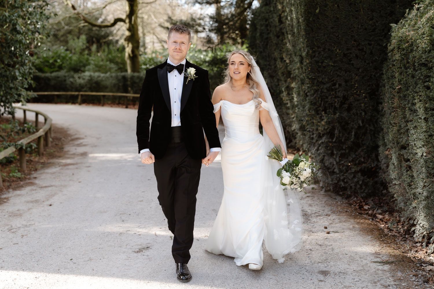 Happy newlyweds strolling together on a tree-lined path, bride holding a white bouquet.