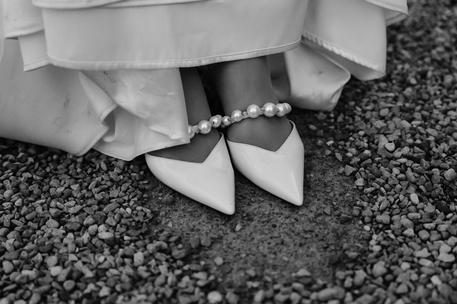 Close-up of bridal white pointed heels adorned with pearl ankle bracelet on gravel ground.