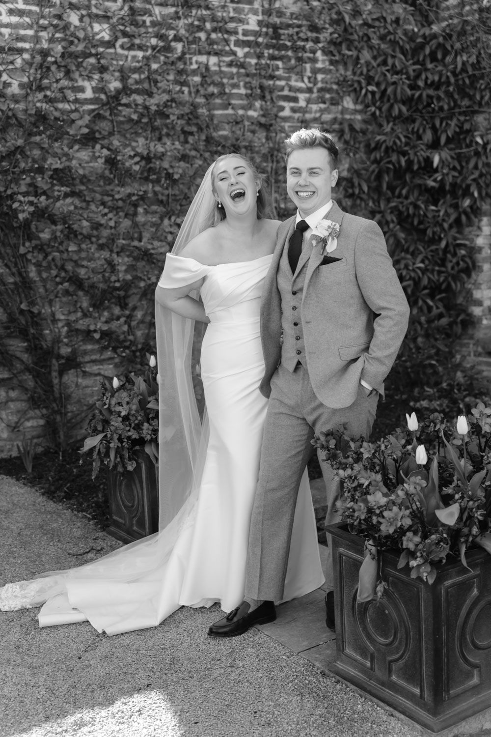 Laughing bride in off-shoulder gown and groom in grey suit posing outdoors.