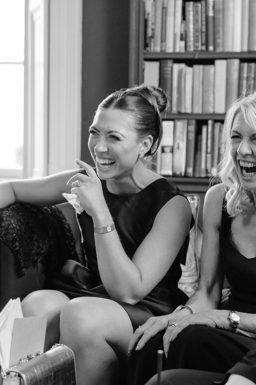 Two women laughing joyfully while seated on a sofa near a bookshelf at a wedding reception.