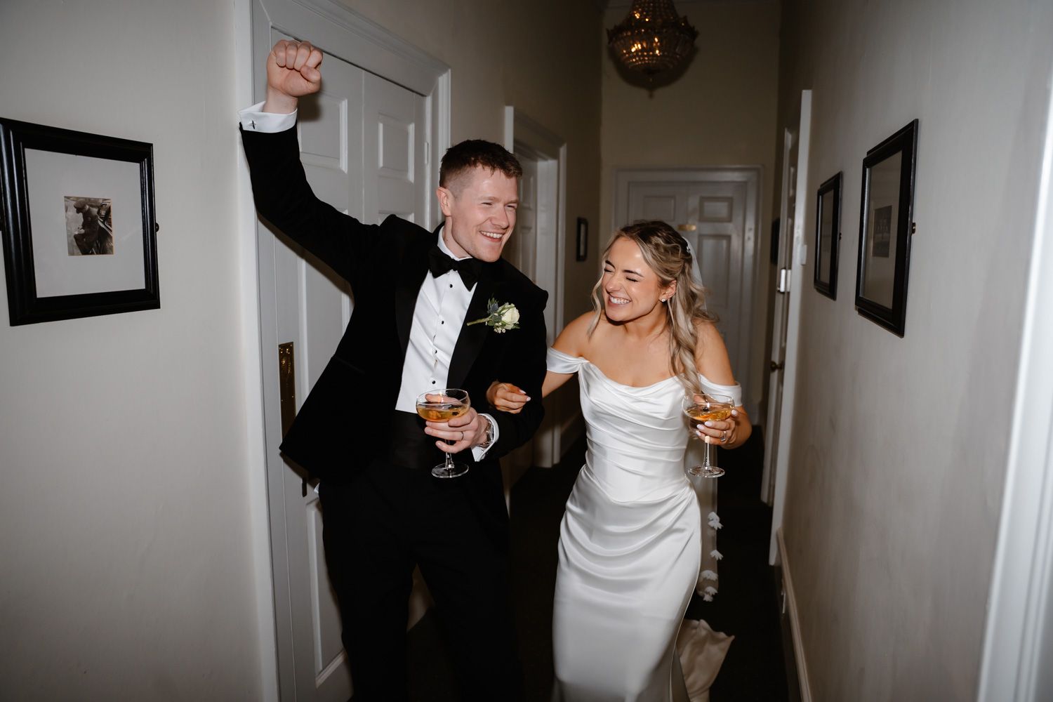 Joyful newlyweds celebrating in hallway holding drinks, groom raising fist.