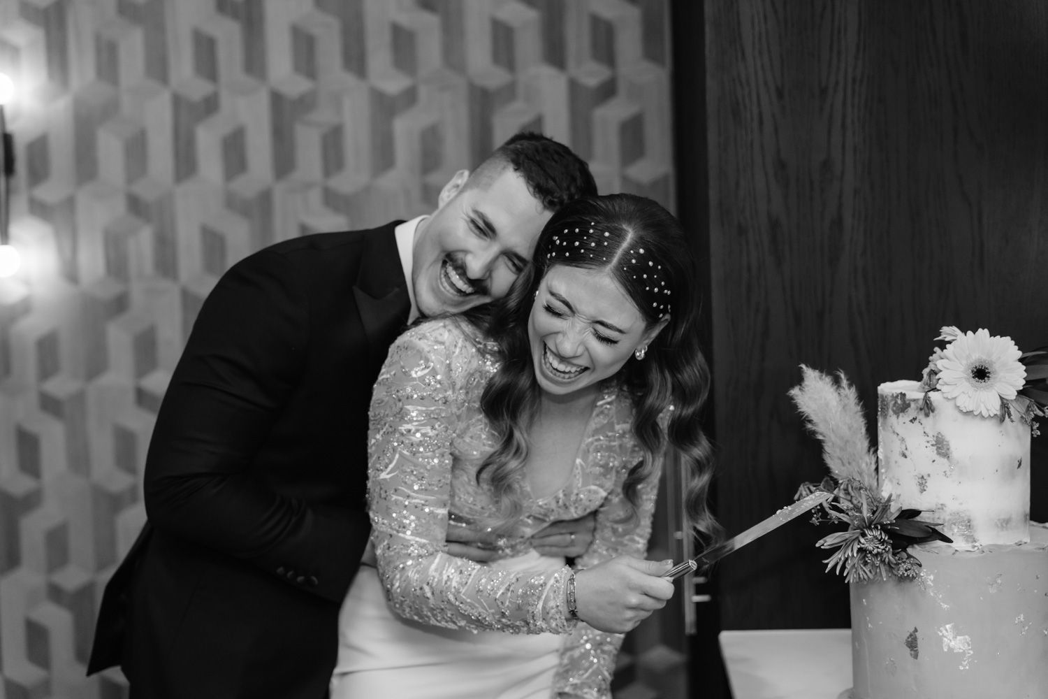 Laughing bride and groom cut wedding cake together in black and white reception photo.