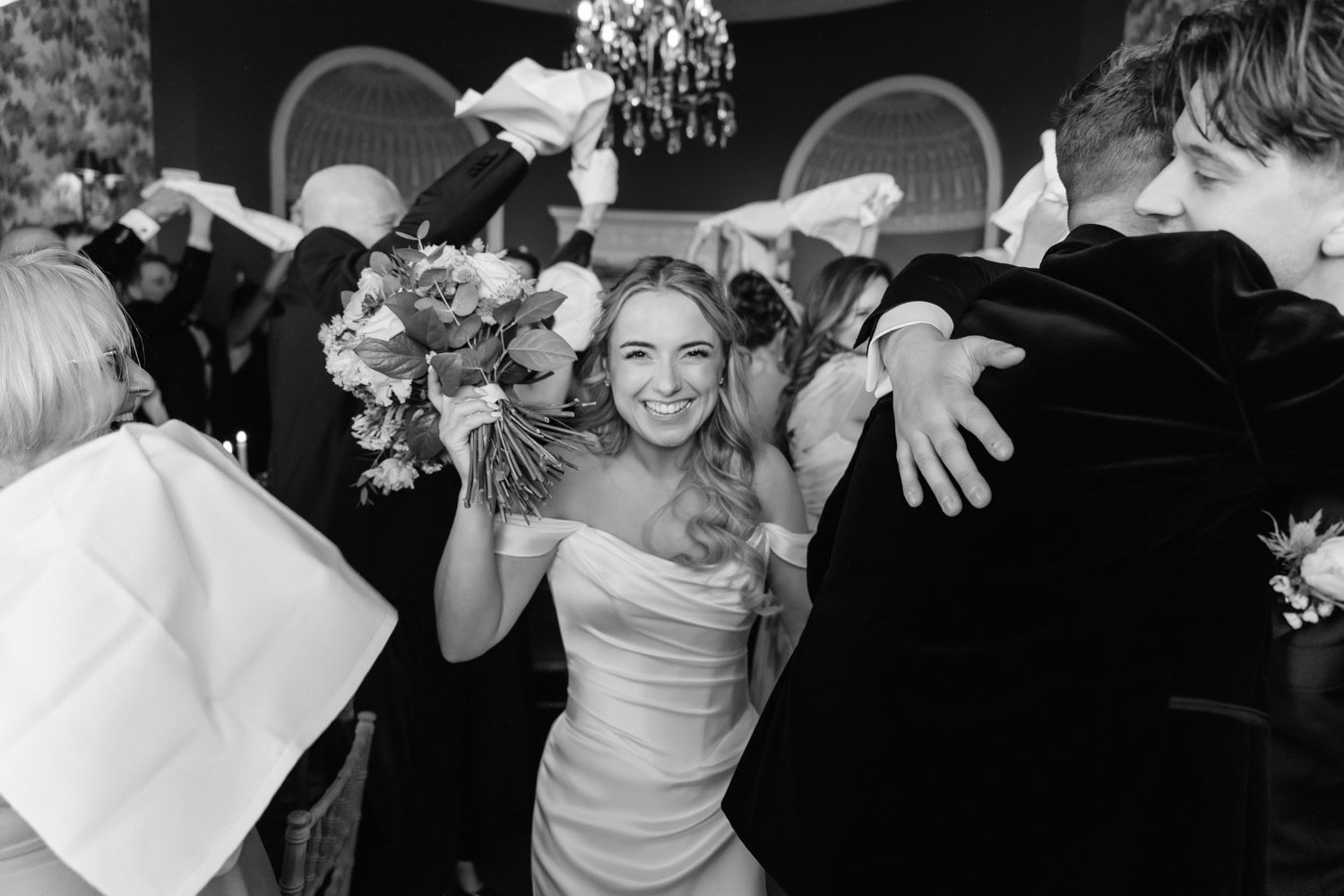 Black and white photo of a smiling bride in a strapless gown holding a bouquet, surrounded by cheering guests at a wedding reception.