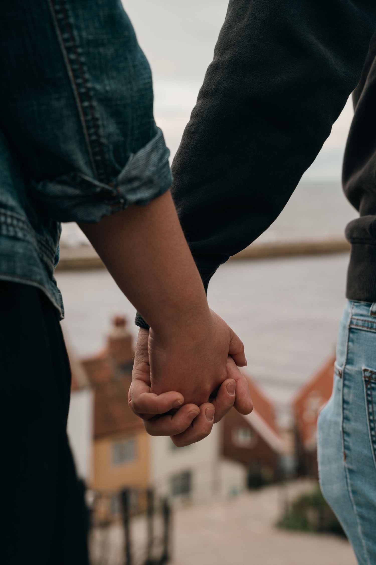 Couple holding hands outdoors near a coastal town, wearing casual denim and dark clothing.