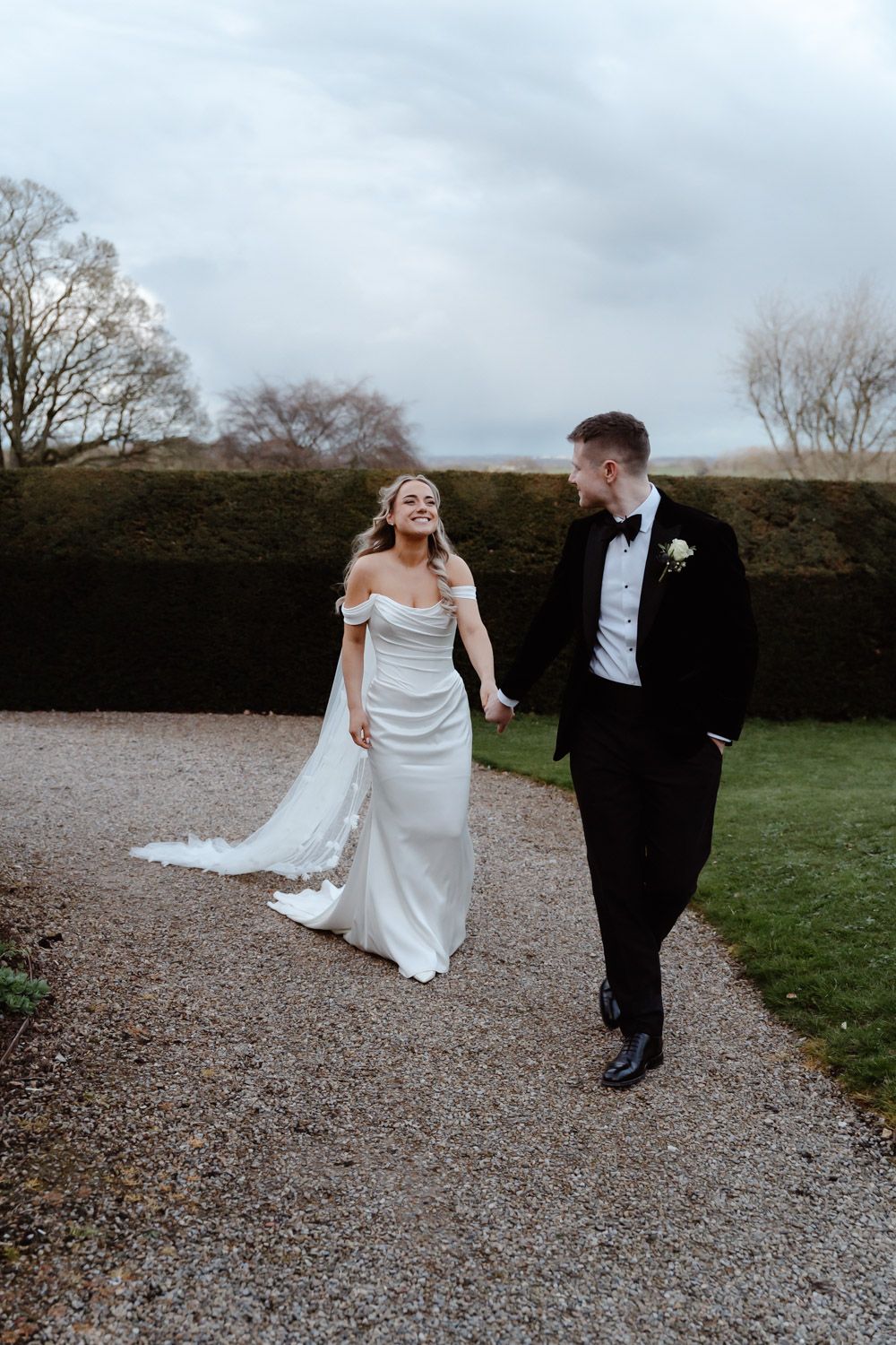 Bride in off-shoulder gown and groom in tuxedo walking hand in hand outdoors.