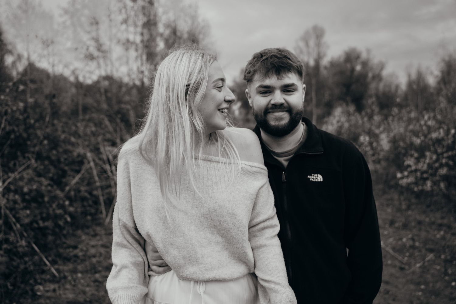 Black and white photo of smiling couple embracing in an autumnal outdoor setting.