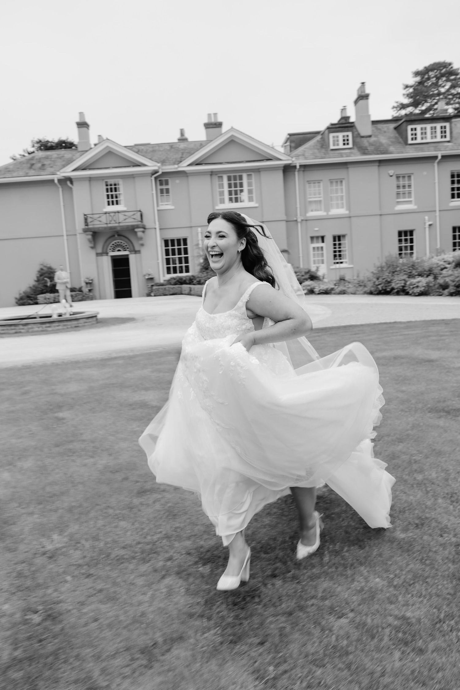 Joyful bride in white gown twirling on lawn before elegant manor house in black and white photo.