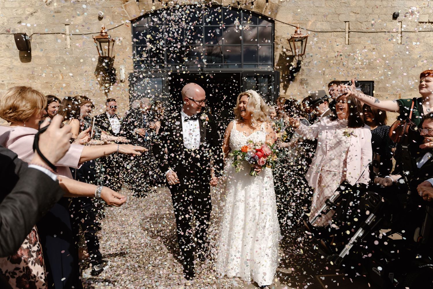 Bride and groom walk through confetti shower outside stone venue surrounded by cheering wedding guests.