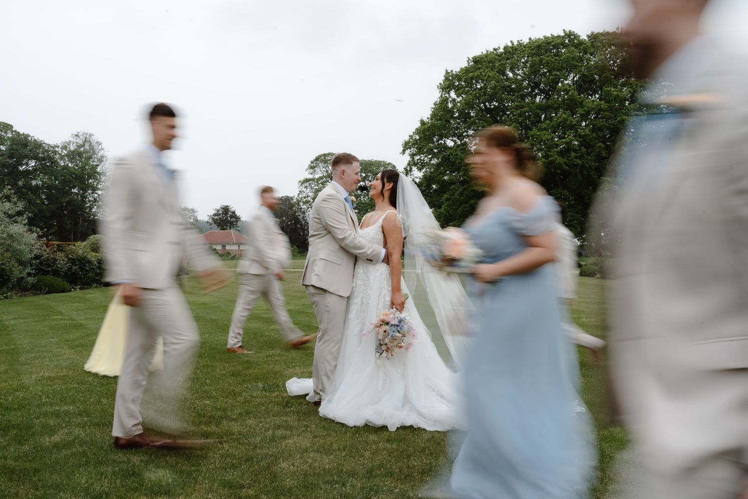 Bride and groom share a kiss outdoors while wedding party members blur past in motion on a green lawn.