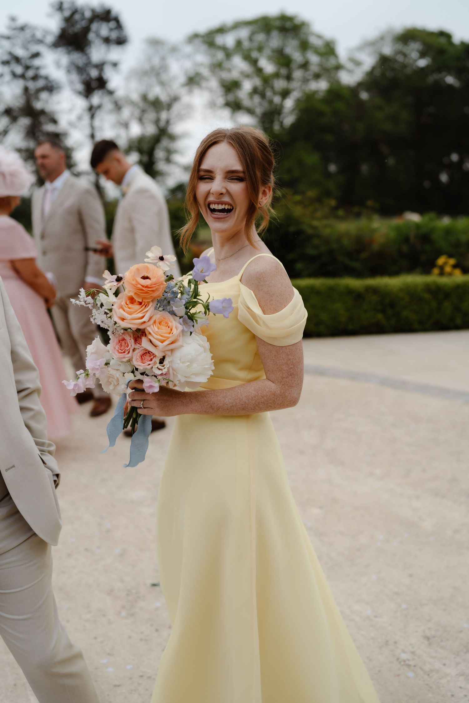 Smiling bridesmaid in yellow gown holds peach and white bouquet at outdoor wedding celebration.