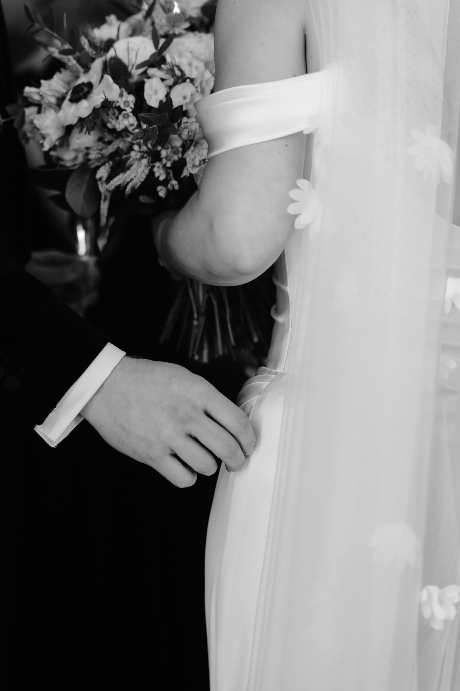 Groom tenderly holding brides hand beside floral veil with daisy appliques in black and white photo.