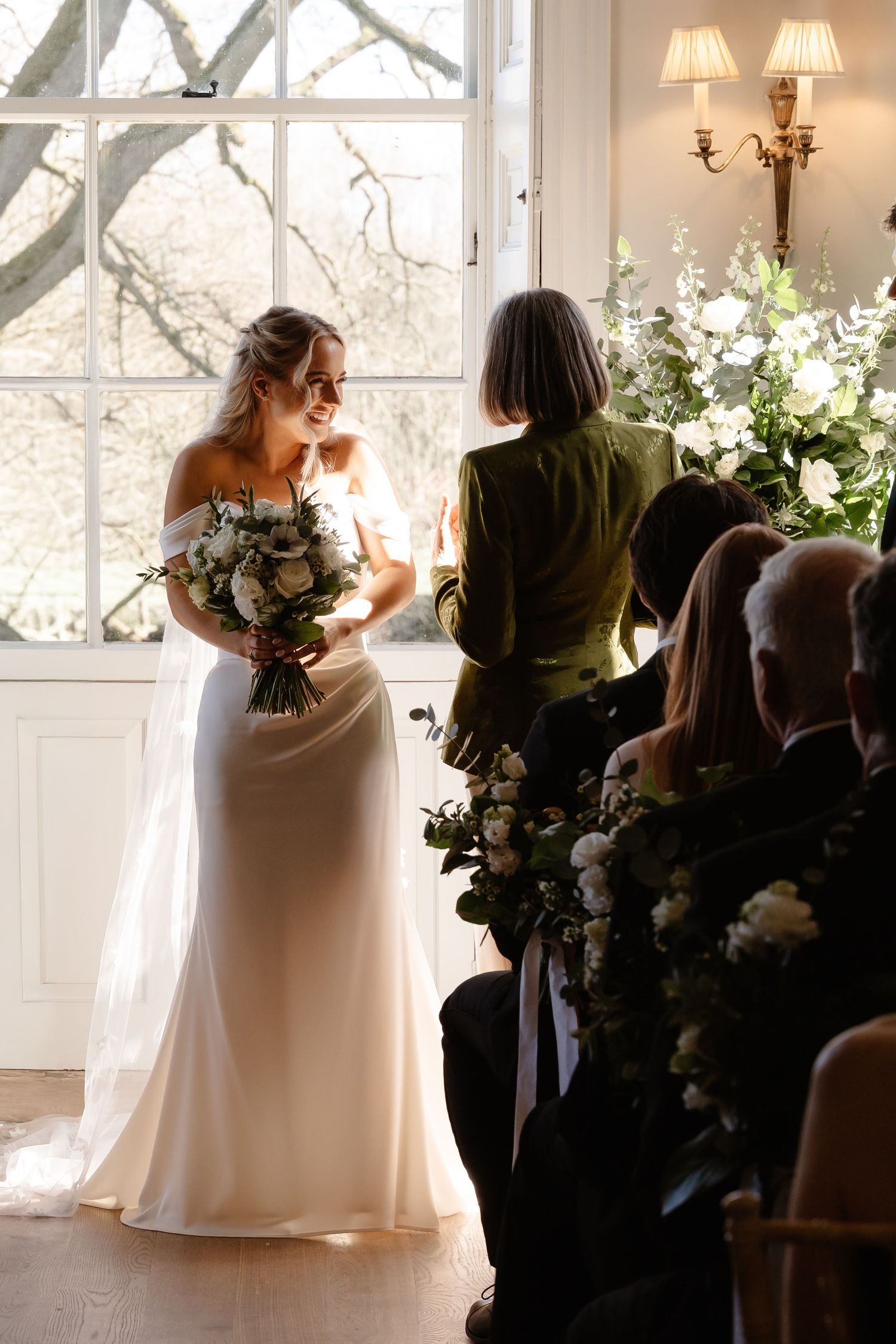 Bride in off-shoulder gown holding bouquet during sunlit indoor wedding ceremony.