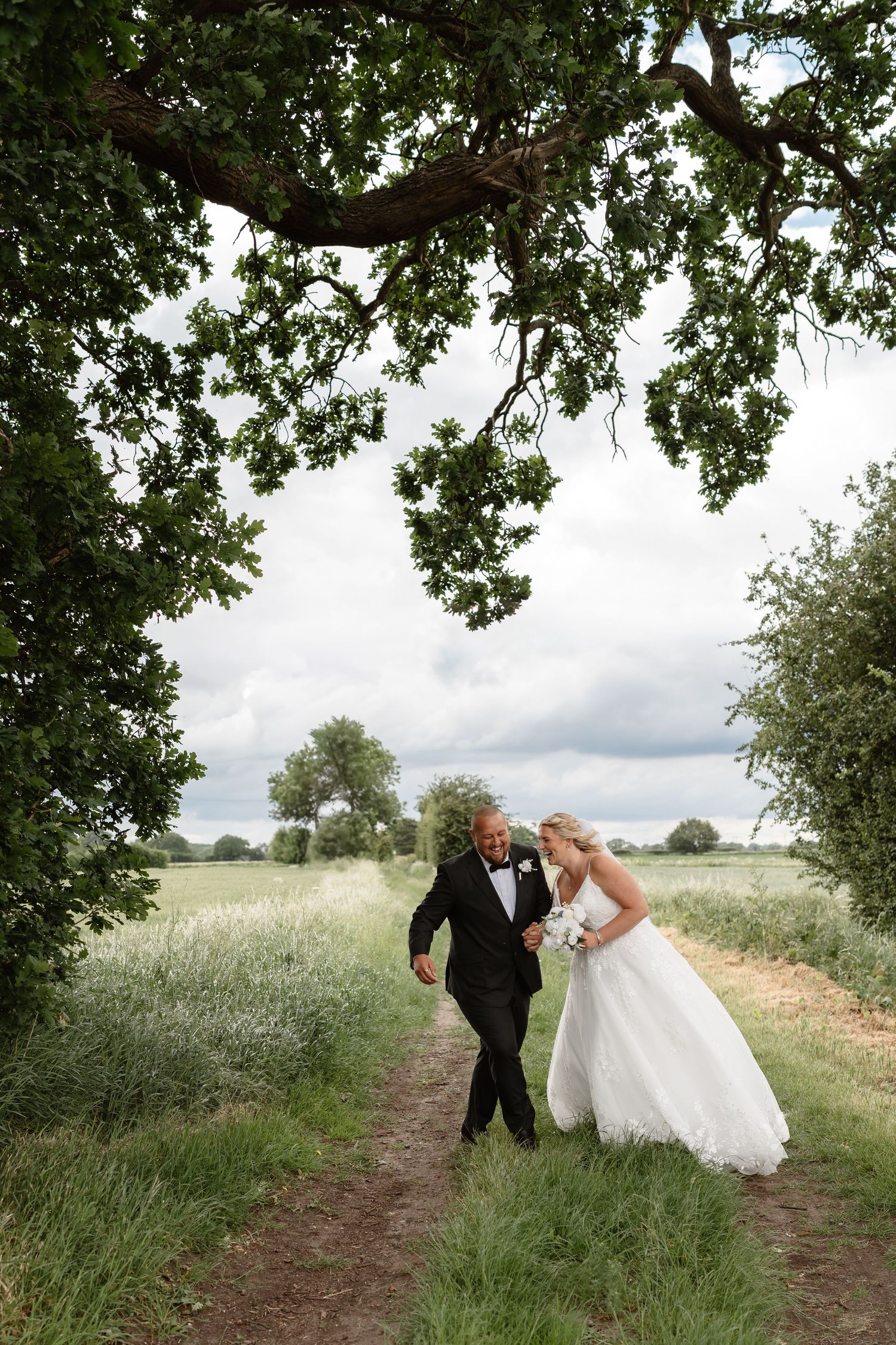 Bride and groom walking hand in hand along a countryside path framed by a large oak tree on their wedding day.