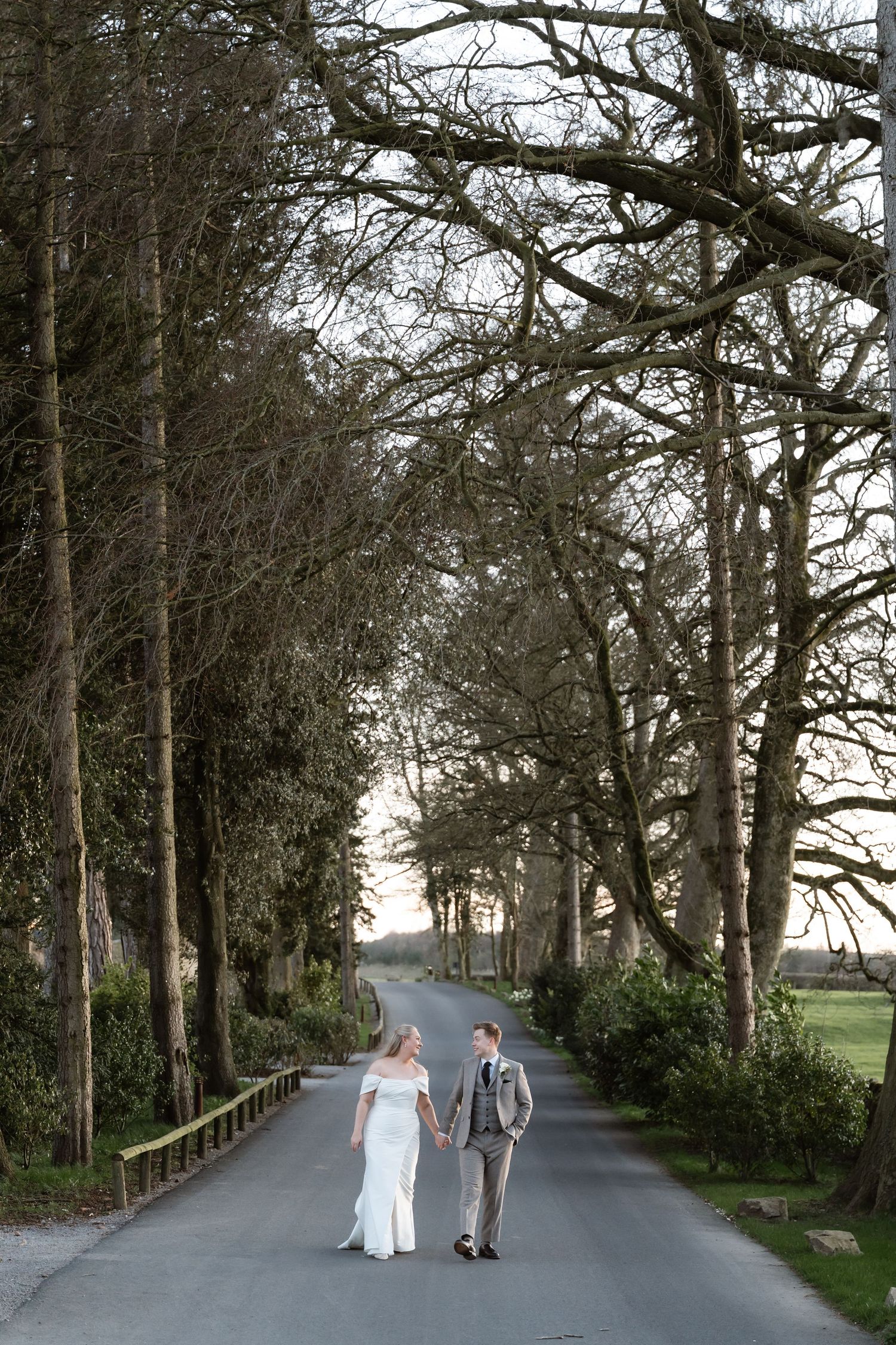 Bride and groom holding hands while walking down a tree-lined avenue at sunset on their wedding day.