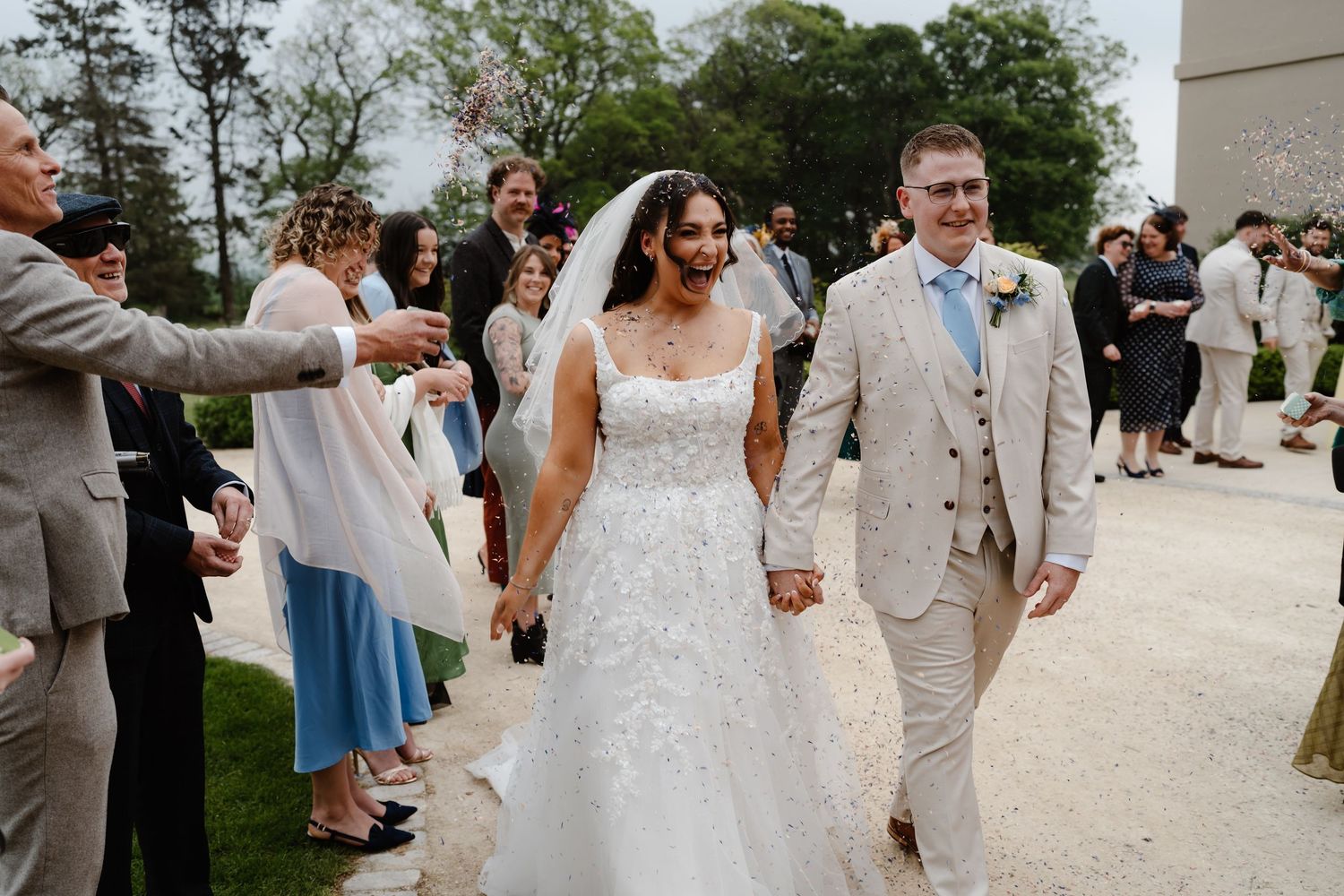 Bride and groom walk hand in hand through cheering wedding guests outdoors on a sunny day.