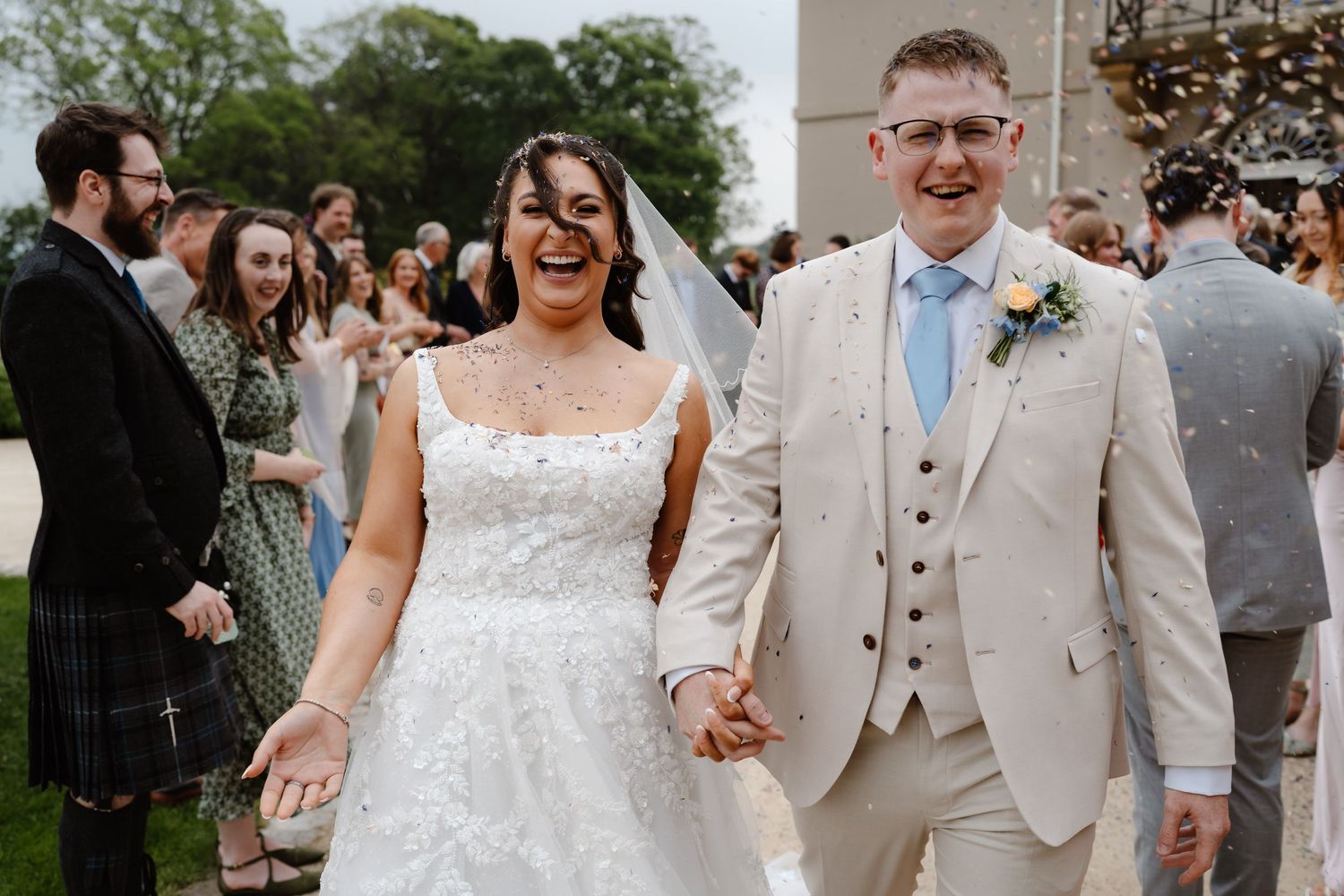 A joyful bride and groom walk hand in hand through confetti at their outdoor wedding ceremony.