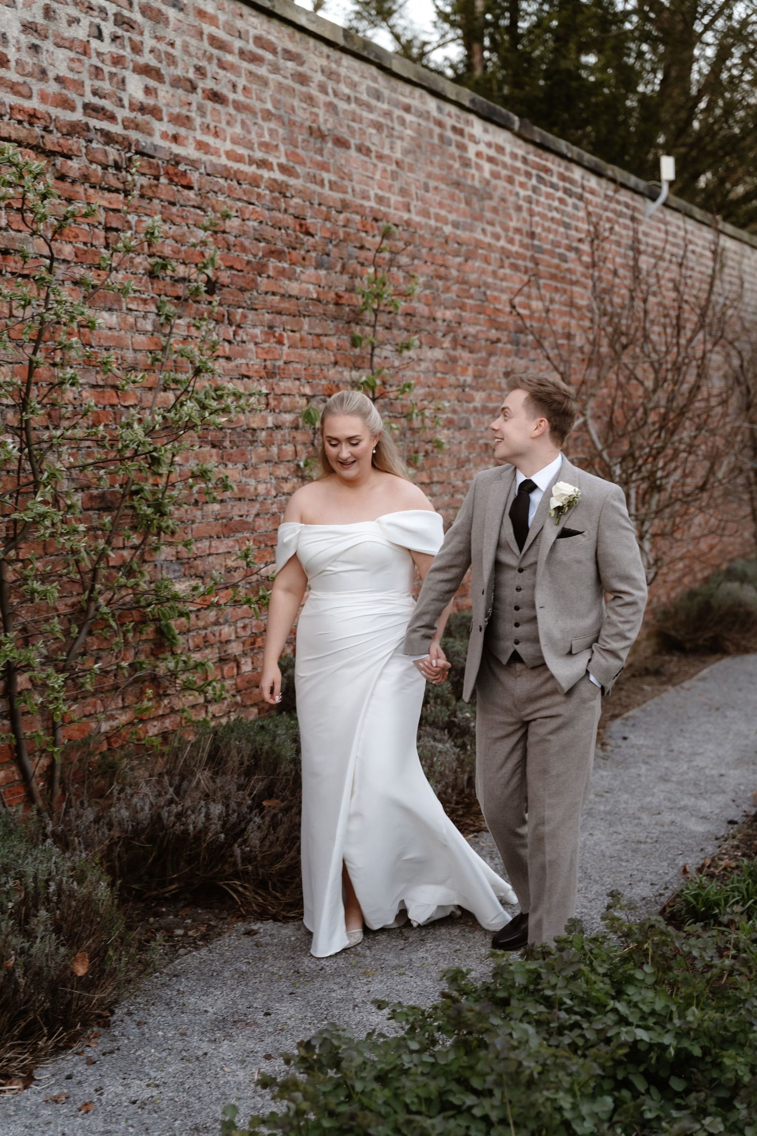 Bride in off-shoulder white gown and groom in grey suit walk hand in hand along a brick wall path.