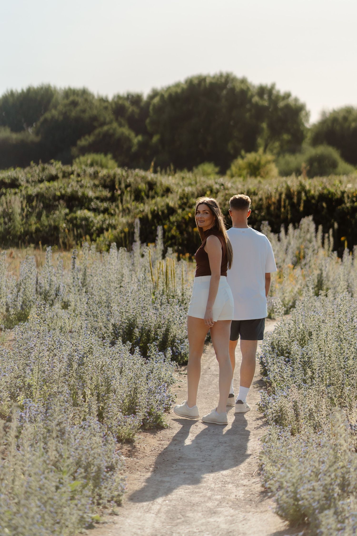 Couple walking hand in hand on a path through white wildflowers with lush green trees in the background.