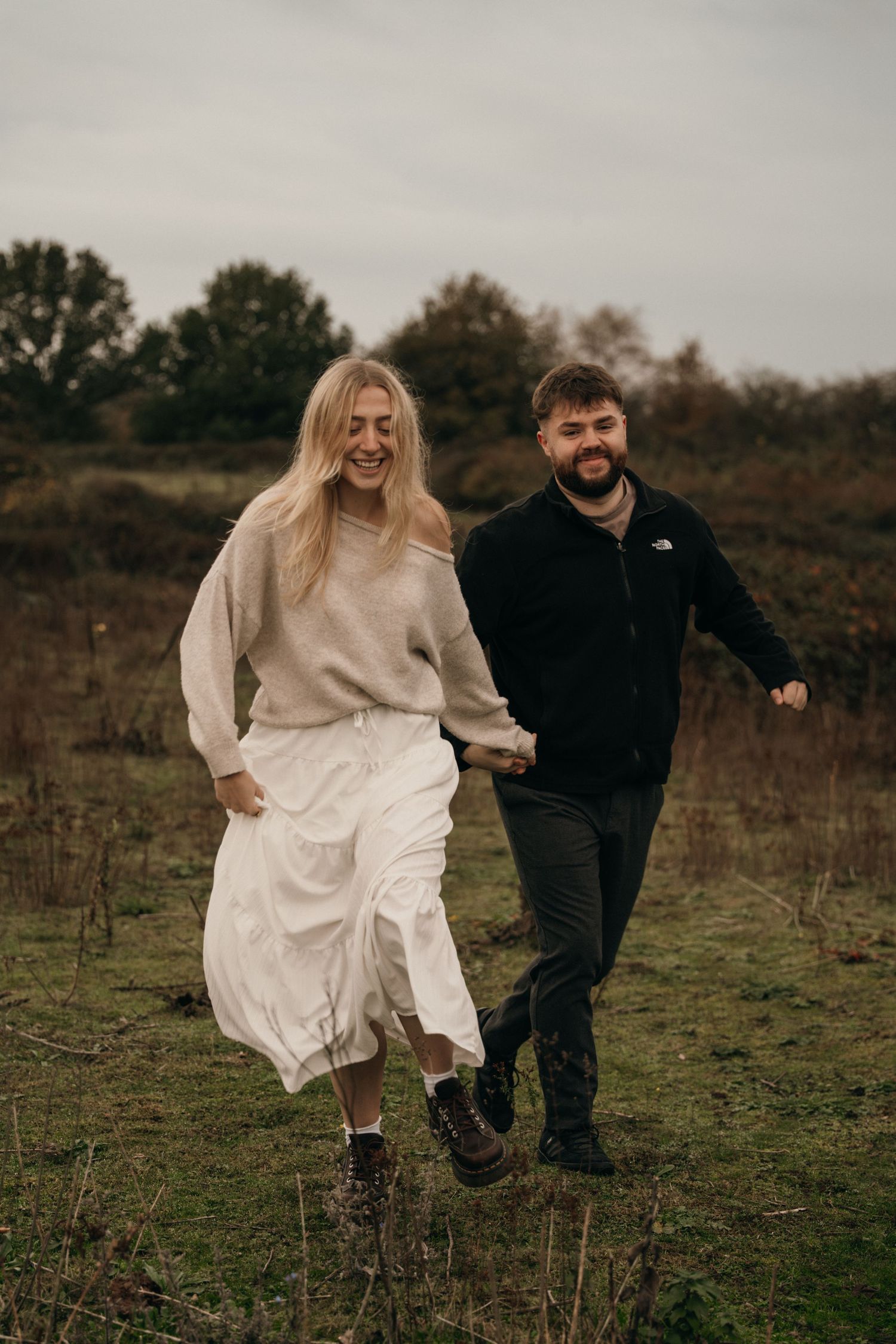 Laughing couple holding hands running through an autumn field, woman in beige sweater and white skirt.