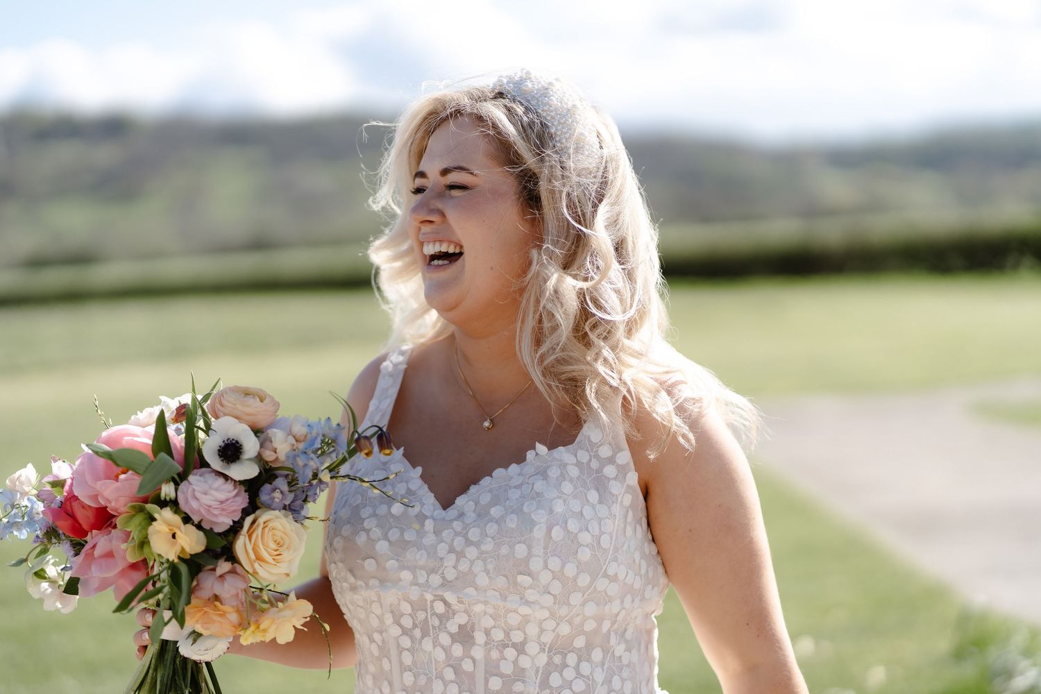Smiling bride in lace wedding dress holding colorful bouquet outdoors on sunny day.
