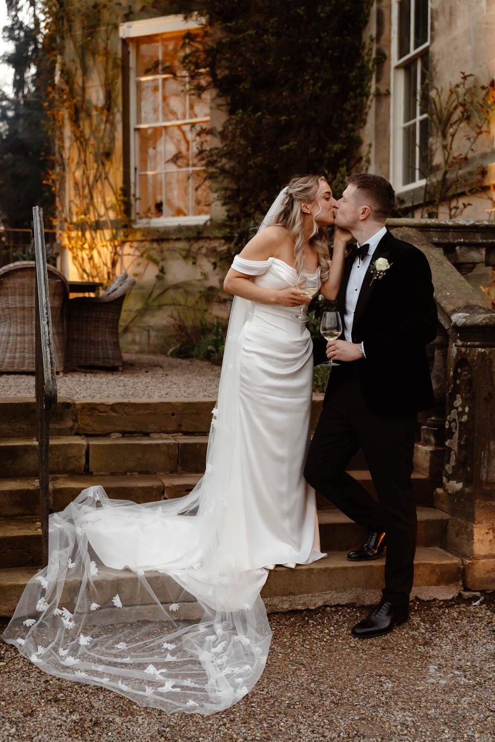 Bride and groom share a kiss on stone steps outside a historic manor, bride in off-shoulder gown with butterfly veil train.