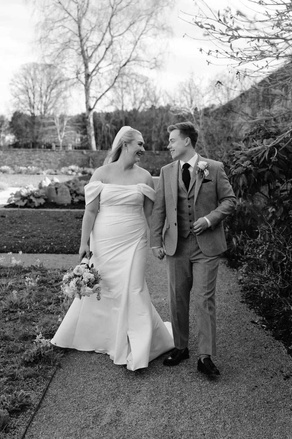 Bride and groom walk hand in hand laughing on a garden path in this elegant black and white wedding photo.