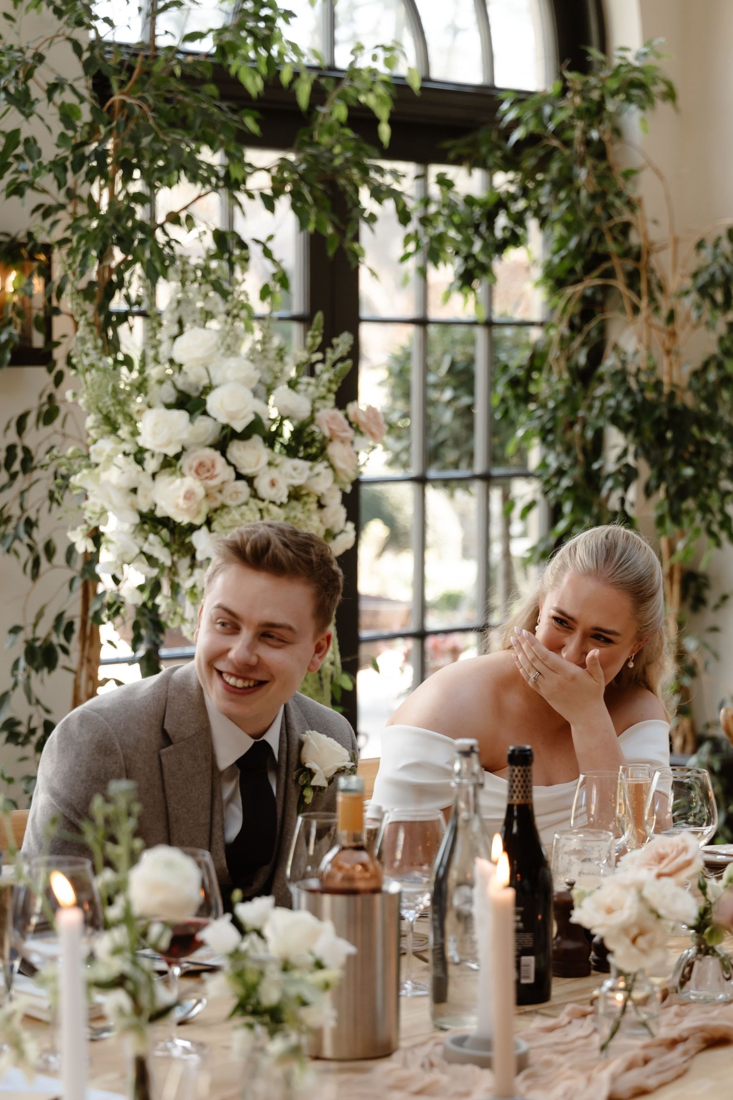 Bride and groom laughing joyfully at wedding reception table adorned with white roses and greenery.