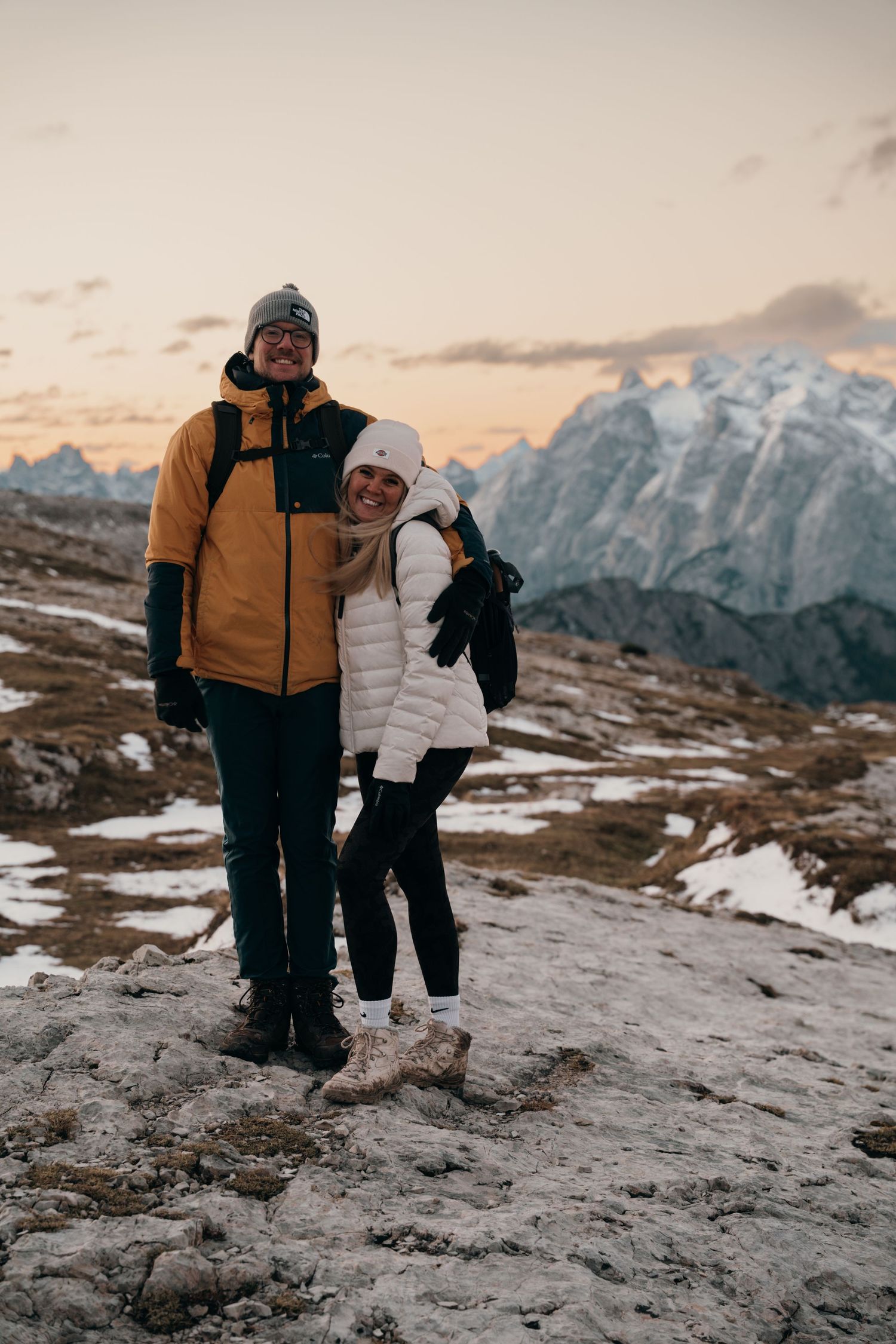 Couple smiling together at golden hour on a rocky alpine trail with snowy peaks behind.