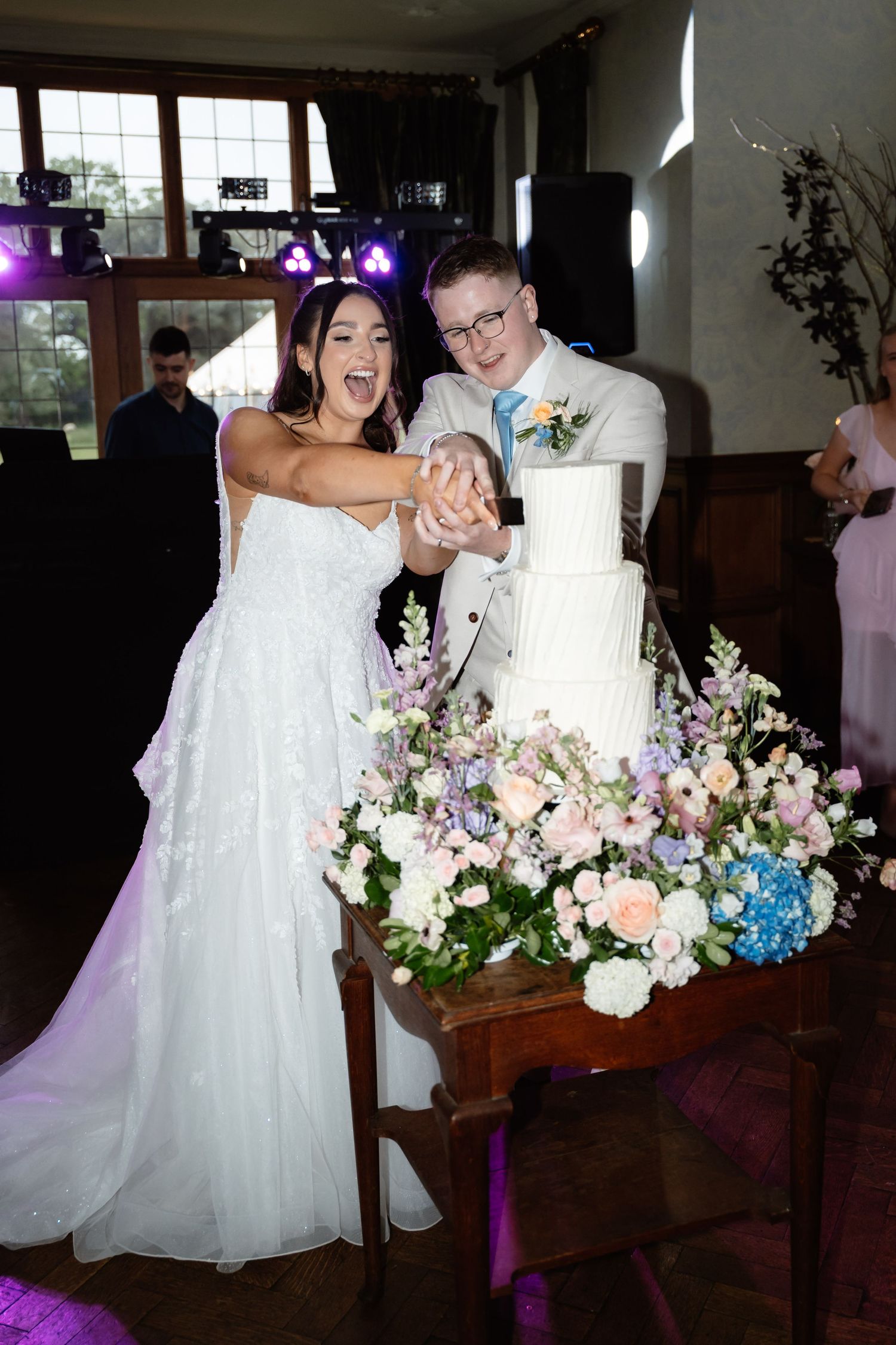 Bride and groom cutting a white tiered wedding cake surrounded by pink and blue floral arrangements.