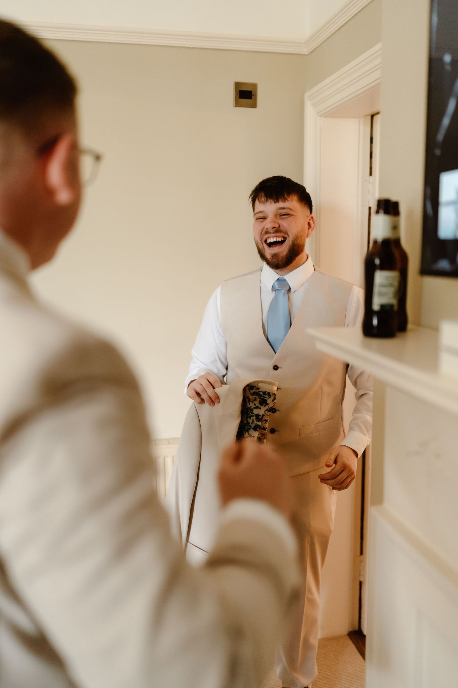 Groom laughing joyfully with groomsman while getting ready in elegant white room on wedding day.
