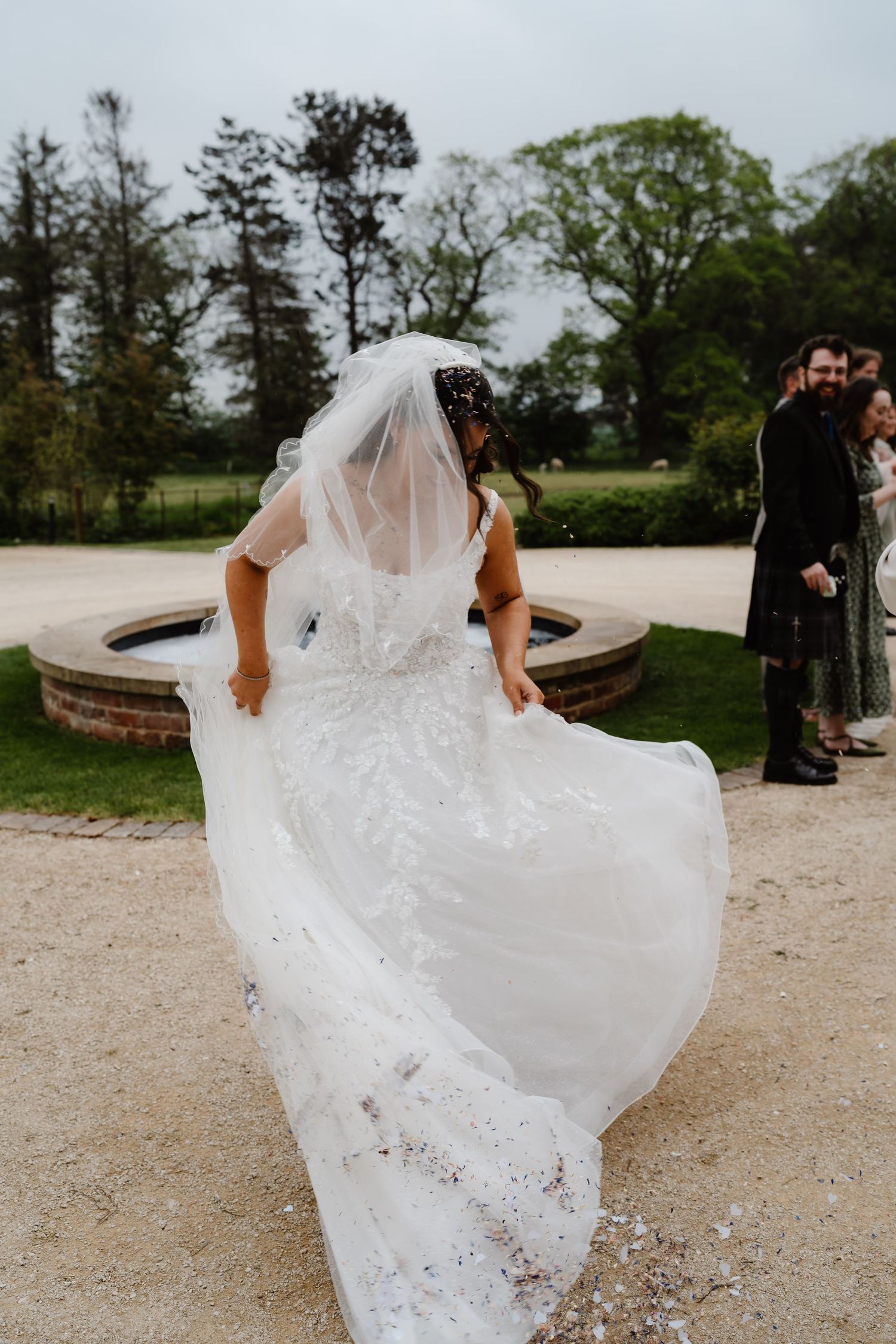 Bride in a beaded white wedding gown and veil twirling outdoors, with confetti falling around her at a garden venue.