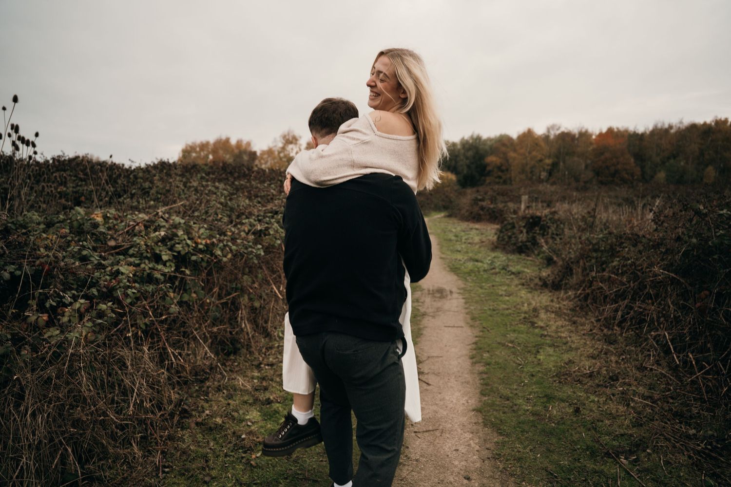 Couple walking along a rural autumn path, woman smiling back while being carried by her partner.