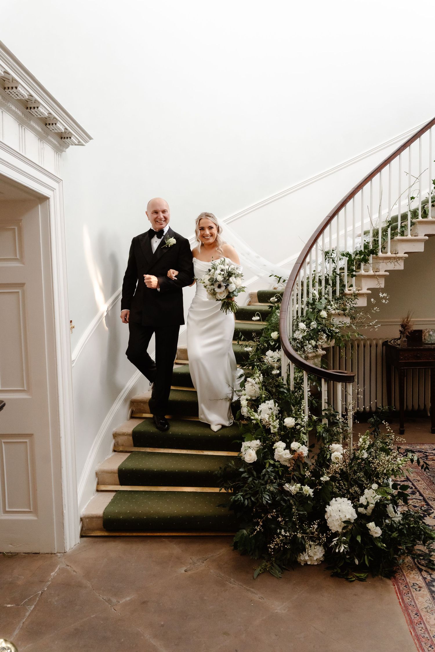 Bride and groom descending floral-adorned staircase decorated with white flowers and greenery.