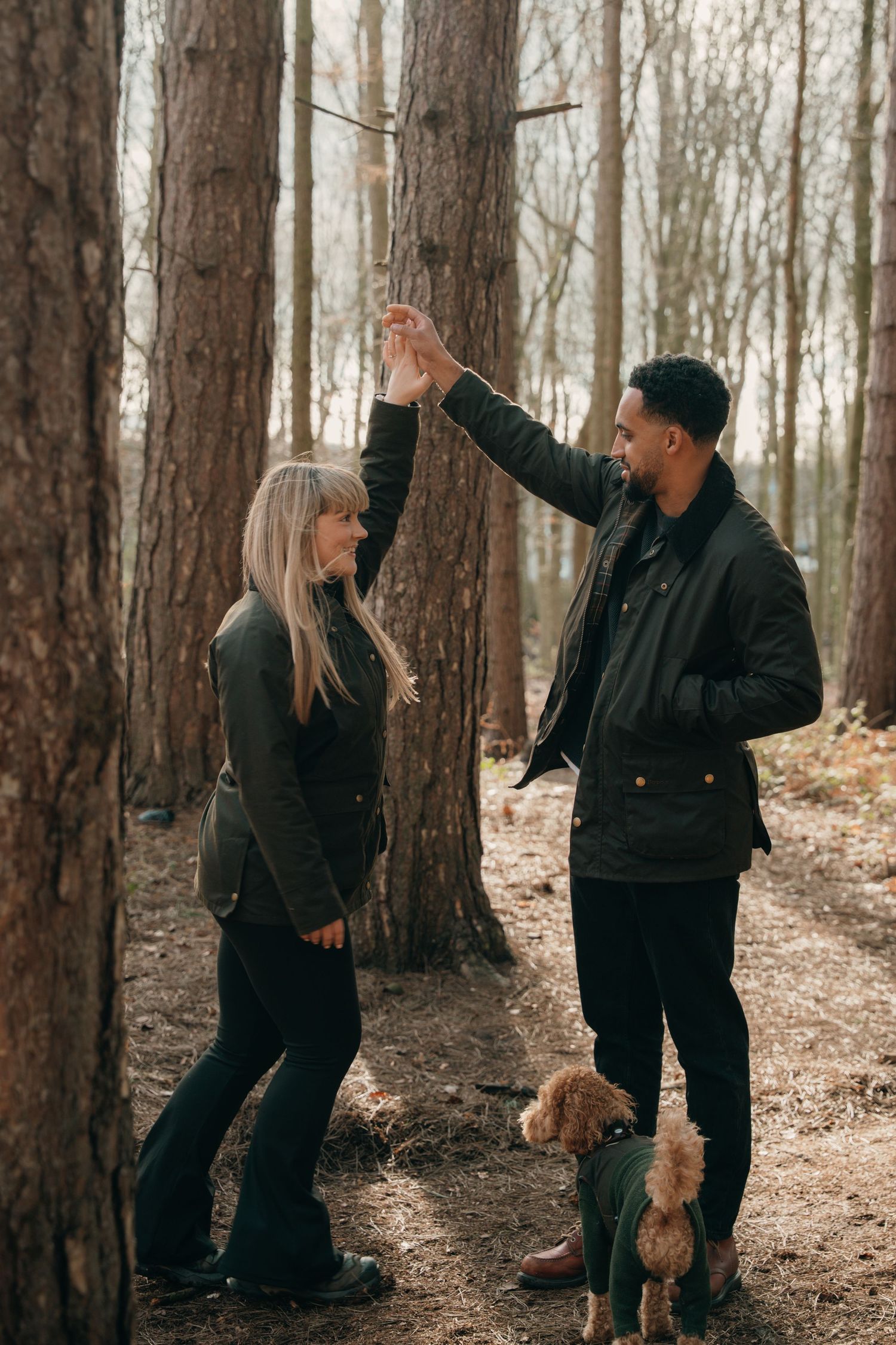 Couple dancing playfully among trees in a forest with their small doodle dog nearby.
