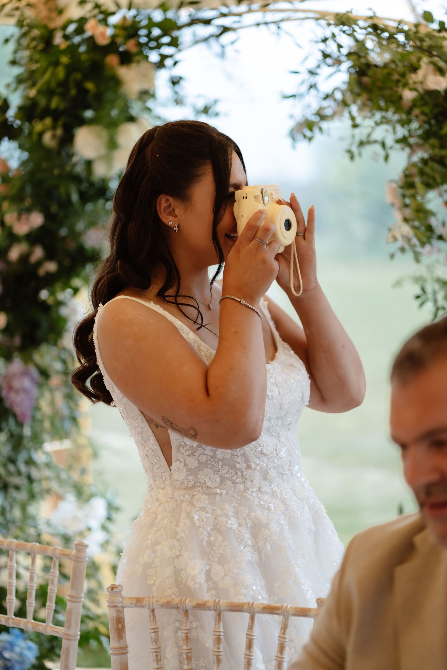 Bride in white lace dress taking photo with Instax camera under floral arch at outdoor wedding ceremony.