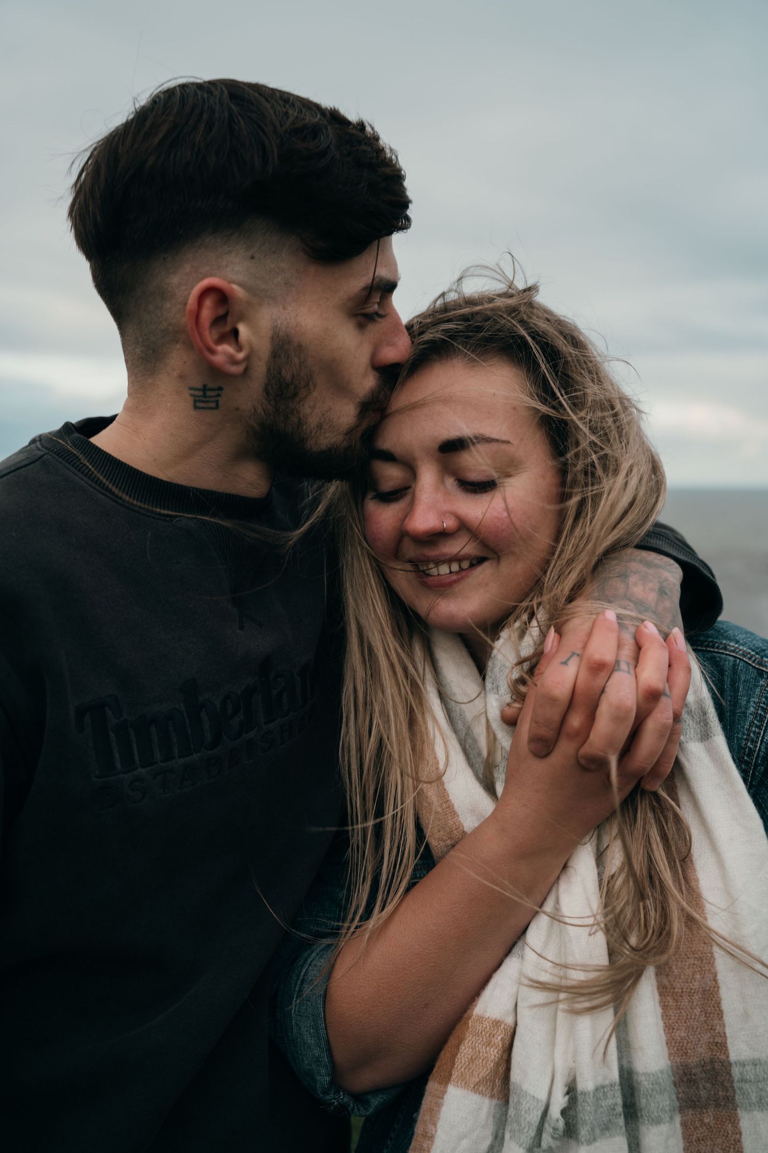 Young couple embracing outdoors, man kissing woman on cheek while she smiles under cloudy skies.