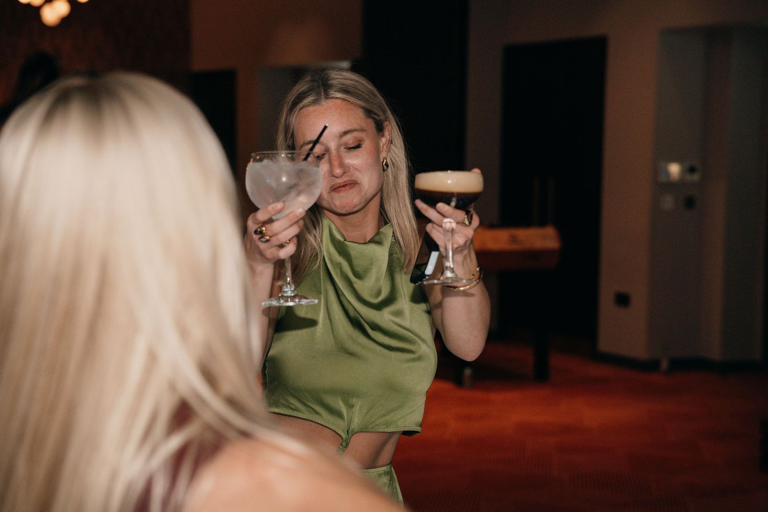 Woman in green dress holding two cocktail glasses at an indoor evening event.