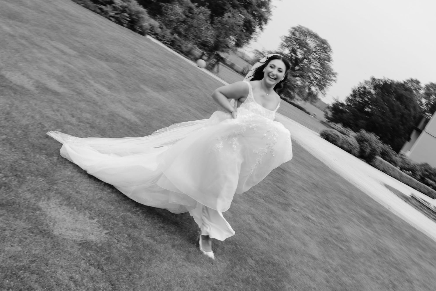 Black and white photo of a bride in a flowing gown twirling joyfully on a lush green lawn.