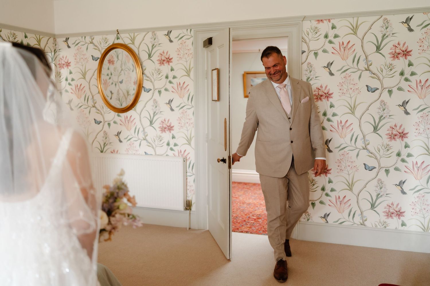Groom in a tan suit smiling as he sees his bride for the first time in a floral wallpapered room.