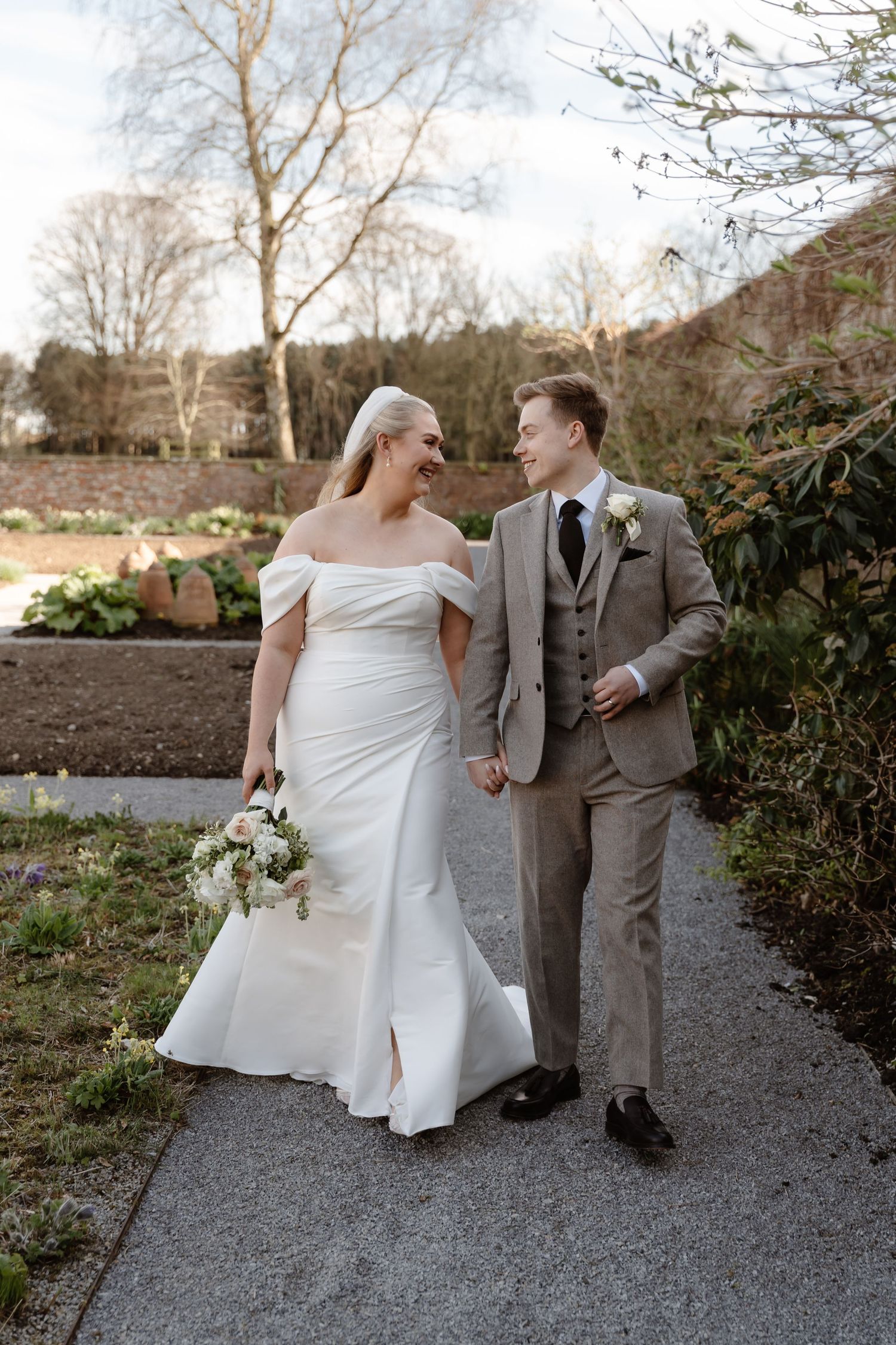 Bride and groom walking hand in hand through garden in wedding attire, bride holding white bouquet.