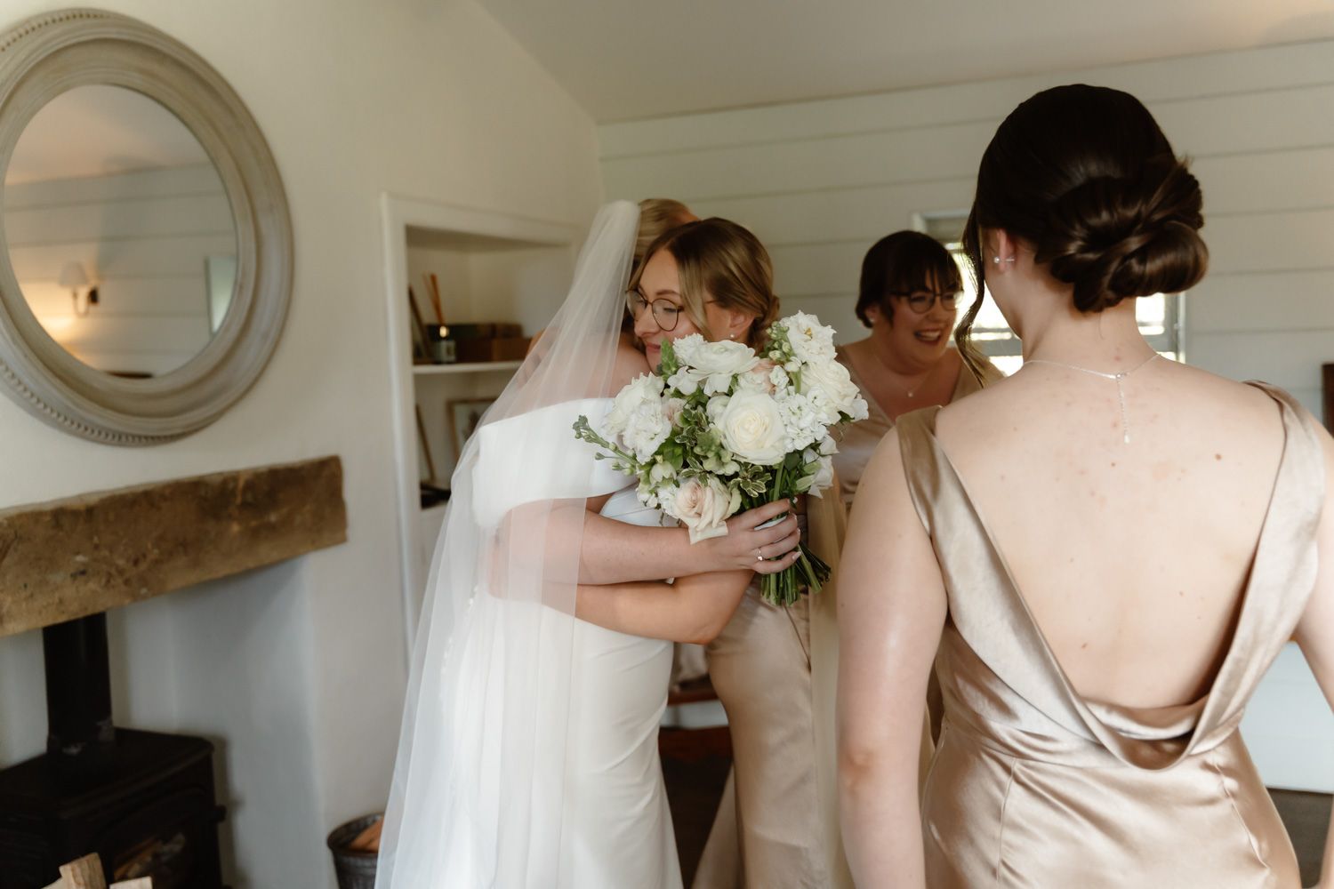Bride embracing bridesmaid while holding white bouquet in rustic wedding preparation room.