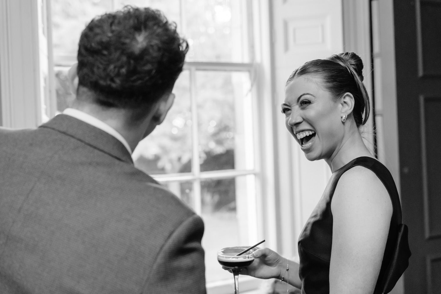 Black and white photo of wedding guest laughing joyfully while holding a drink at reception.