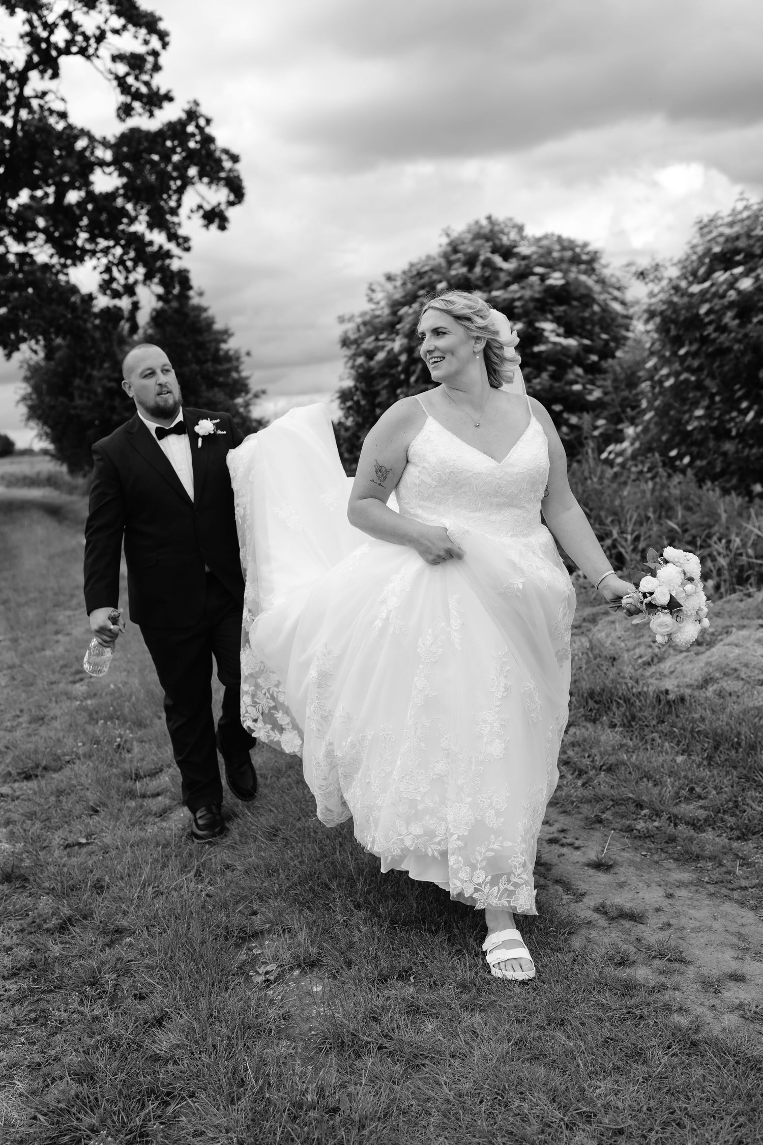 Black and white photo of a smiling bride in a lace gown and groom in a tuxedo walking outdoors on their wedding day.