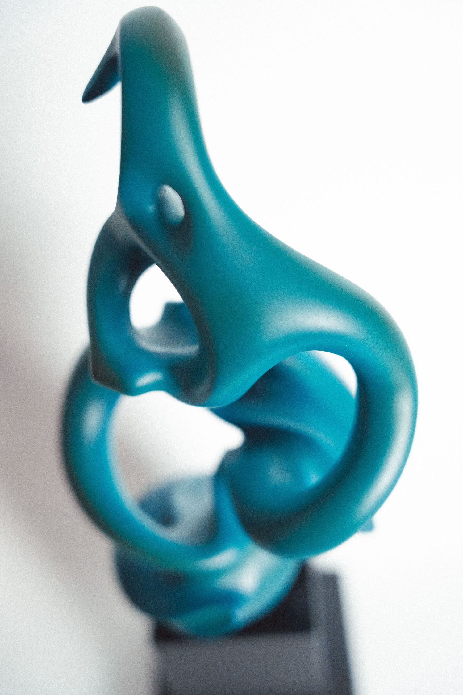 Abstract teal sculpture with flowing curves and organic shapes mounted on a dark base against white background.