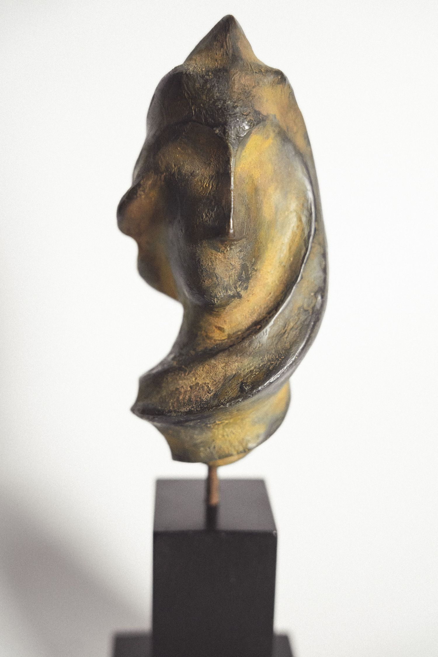 Abstract bronze sculpture of a stylized face profile mounted on a black pedestal, with golden patina finish.