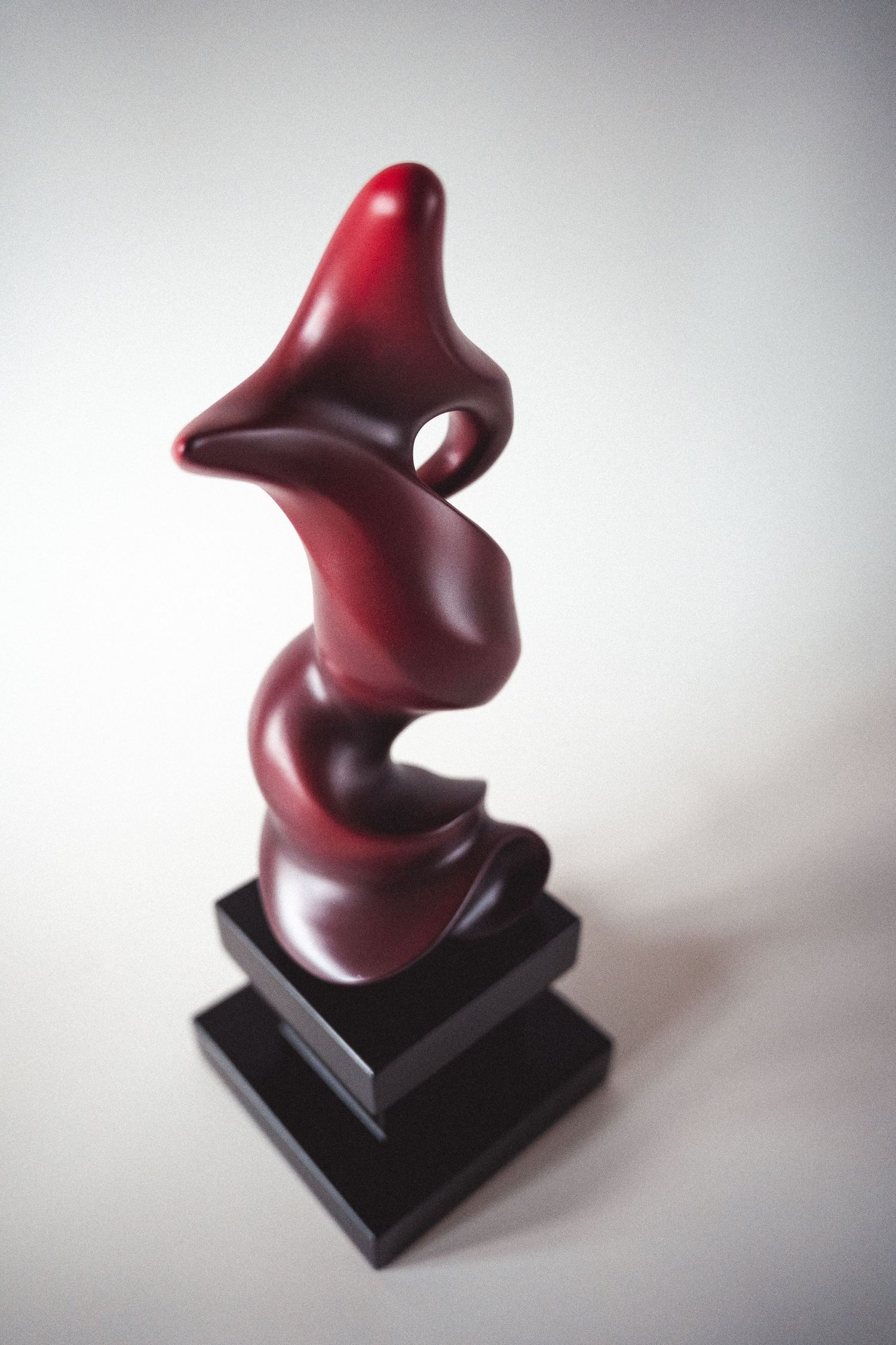 Dark crimson abstract sculpture with spiral organic shape on a black pedestal.