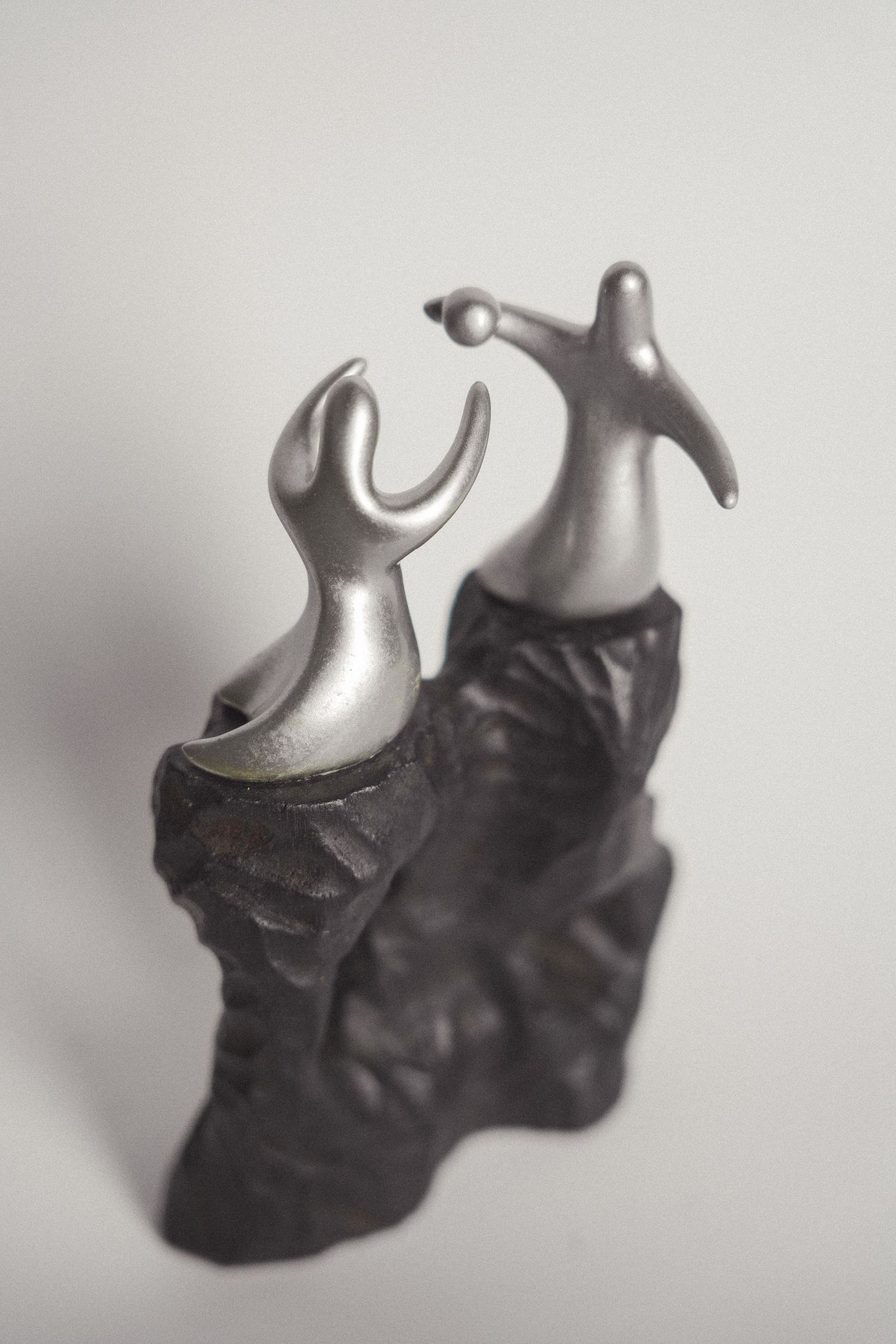 Two abstract silver figurines on dark rocky bases appear to interact, one holding a sphere, in a sculptural art piece.