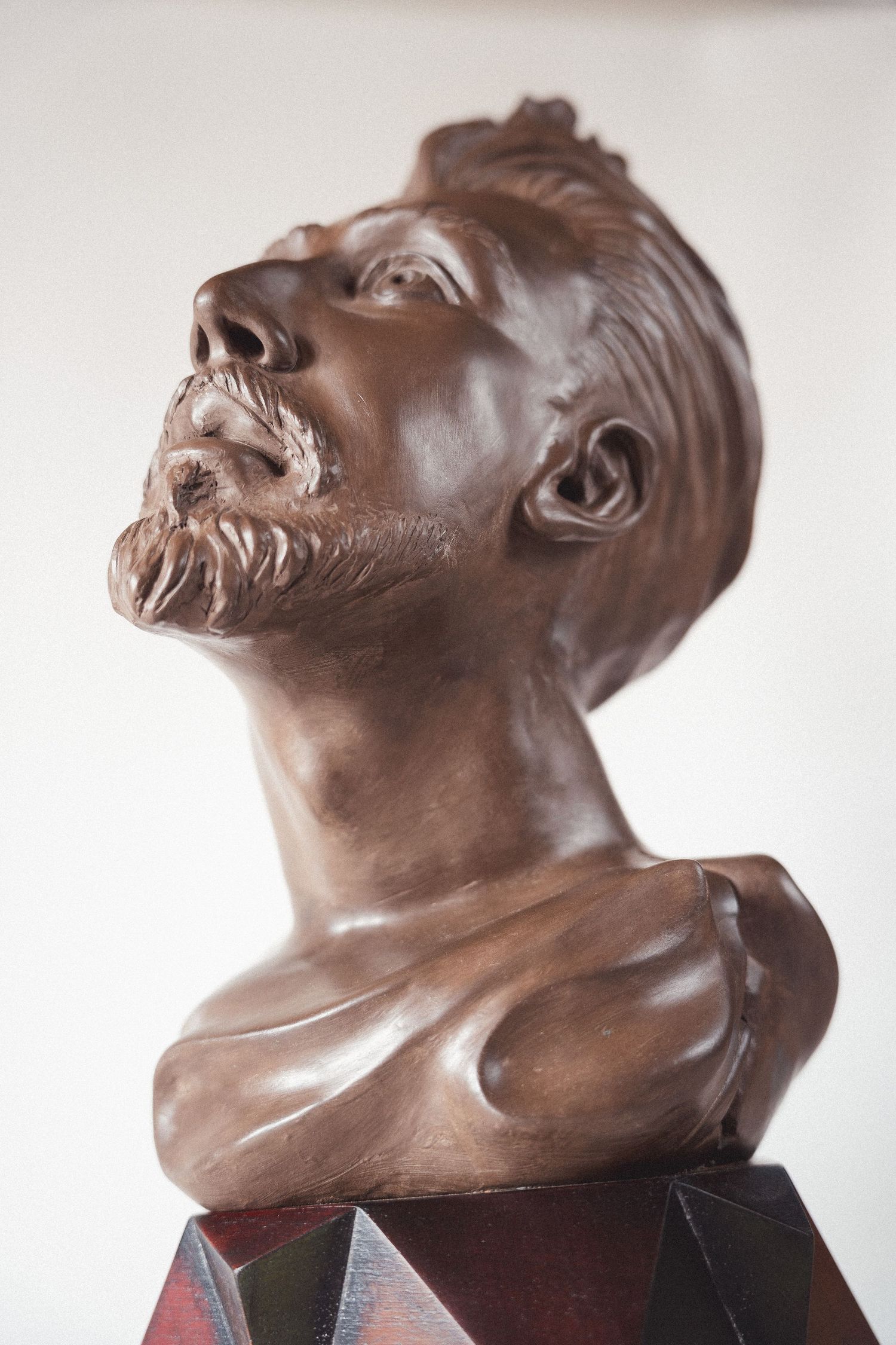 Bronze bust sculpture of a bearded man gazing upward, mounted on a geometric multi-faceted base.