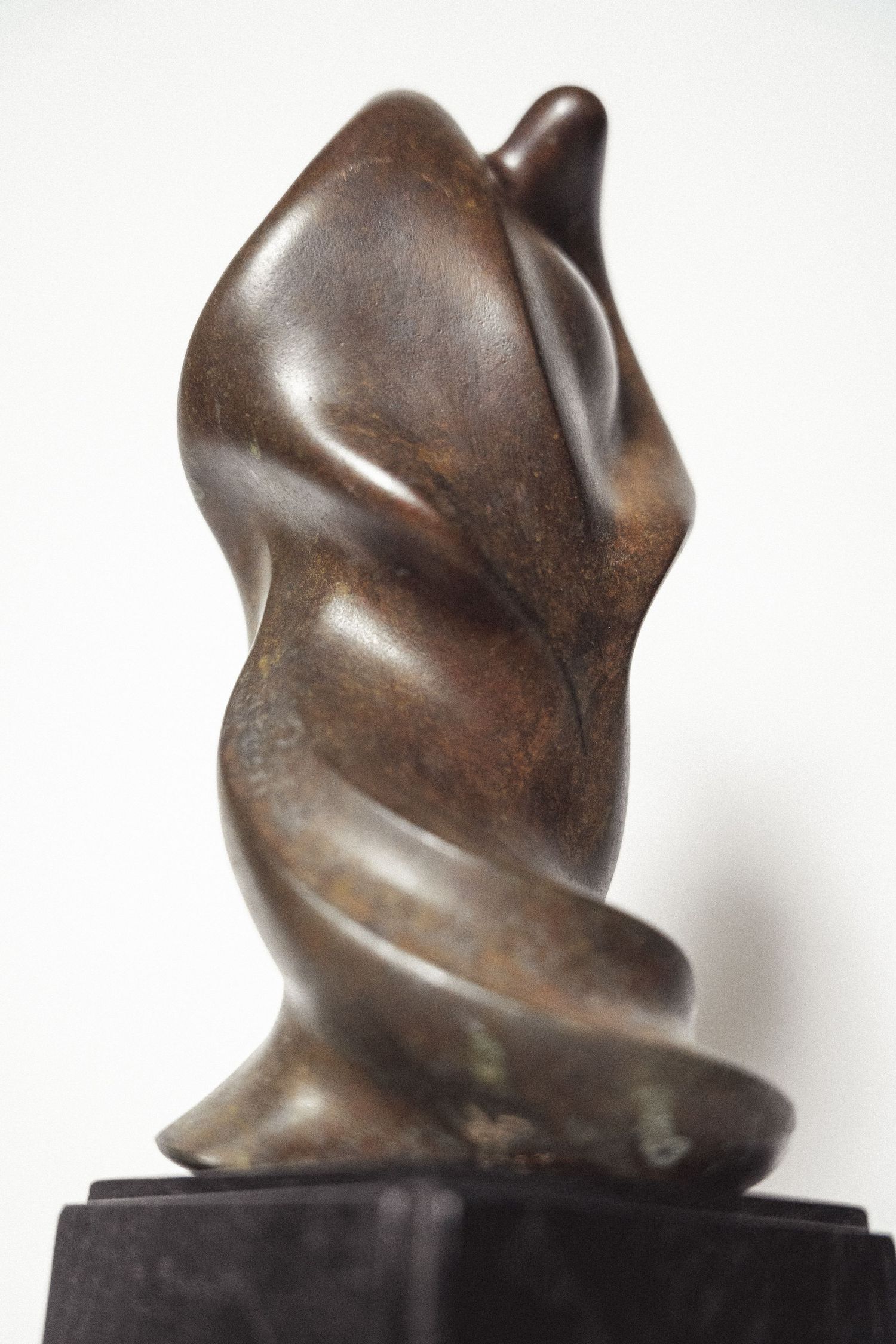Side view of a dark patinated bronze abstract sculpture with twisting organic form on black base.